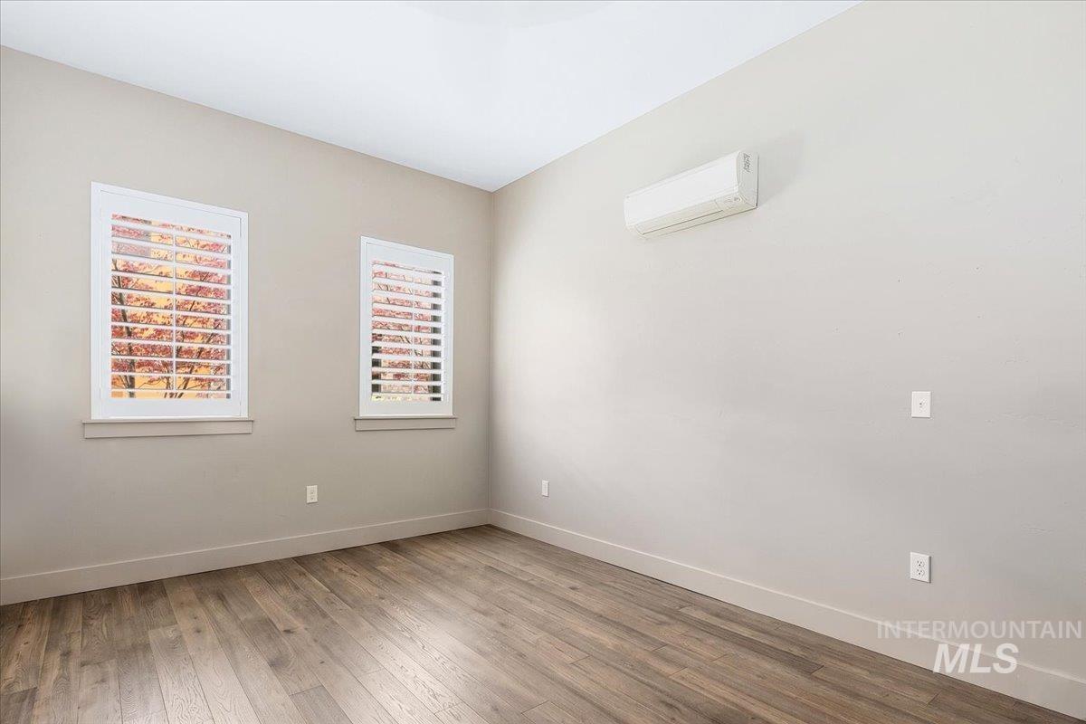 Neutral-toned interior room featuring two windows with white plantation shutters, wood-finish flooring, a wall-mounted air conditioning unit, and white baseboards