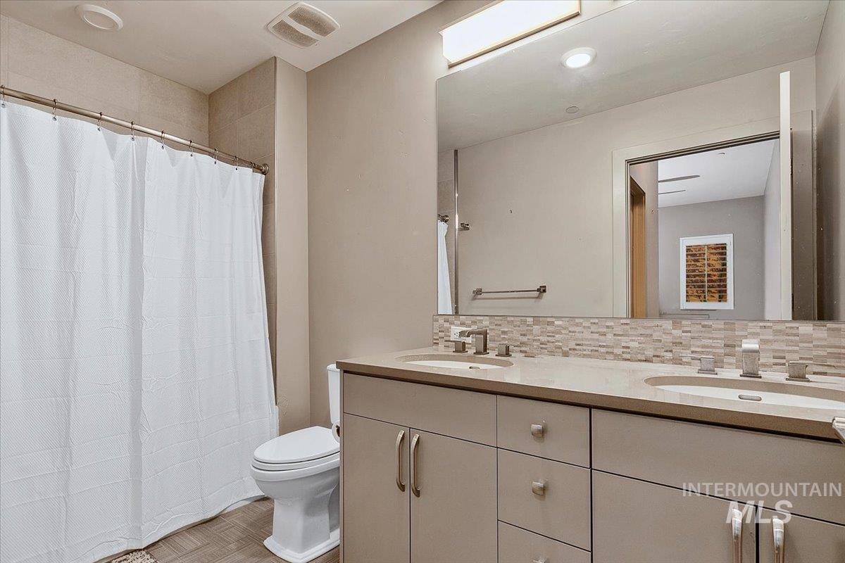 Bathroom featuring a dual vanity with light-toned countertops, contemporary cabinetry, a tile backsplash, and a shower with a tiled surround
