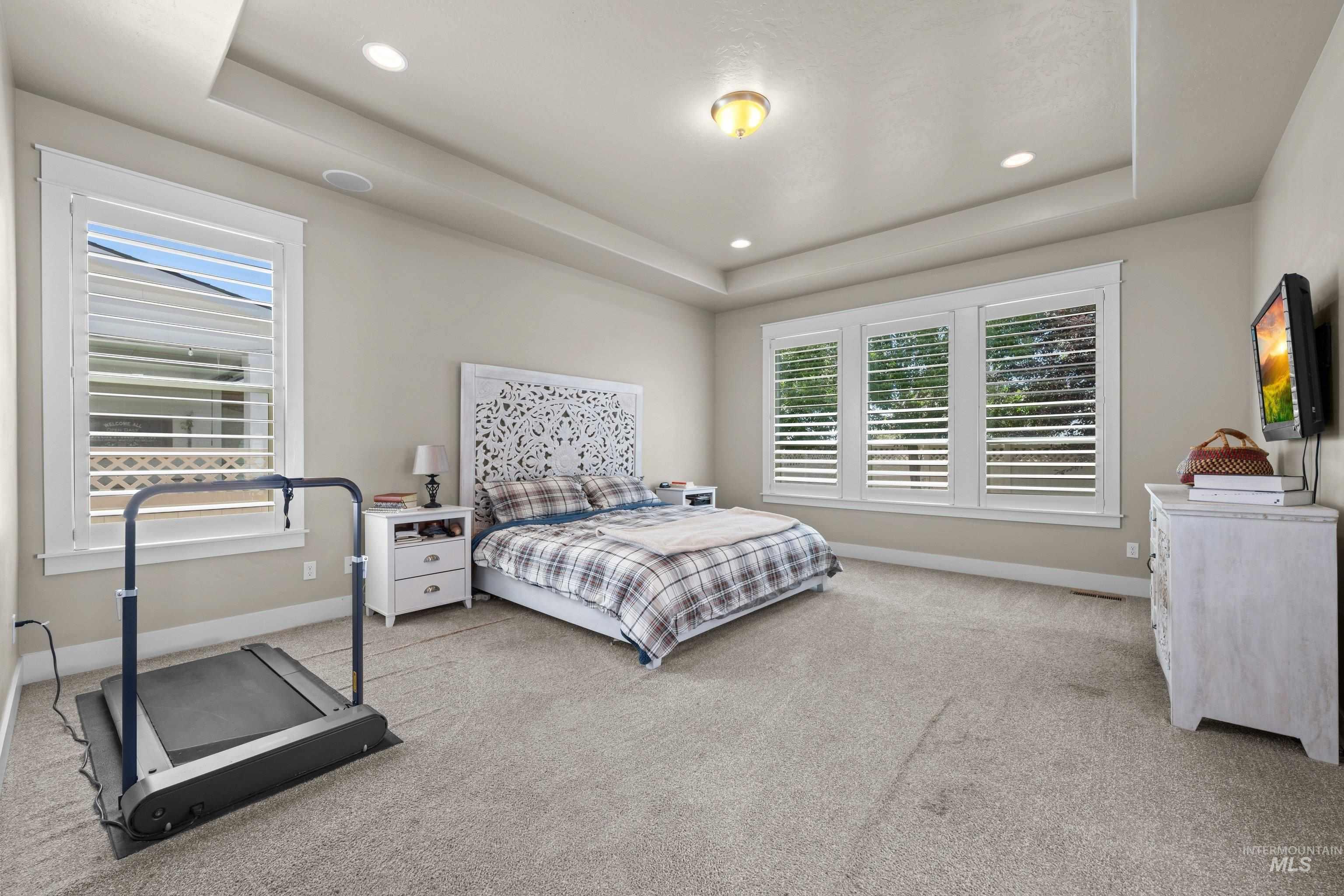 Recessed lighting illuminates this room featuring tray ceilings, a trio of windows with plantation shutters, a single window with plantation shutters, and light-toned carpeting