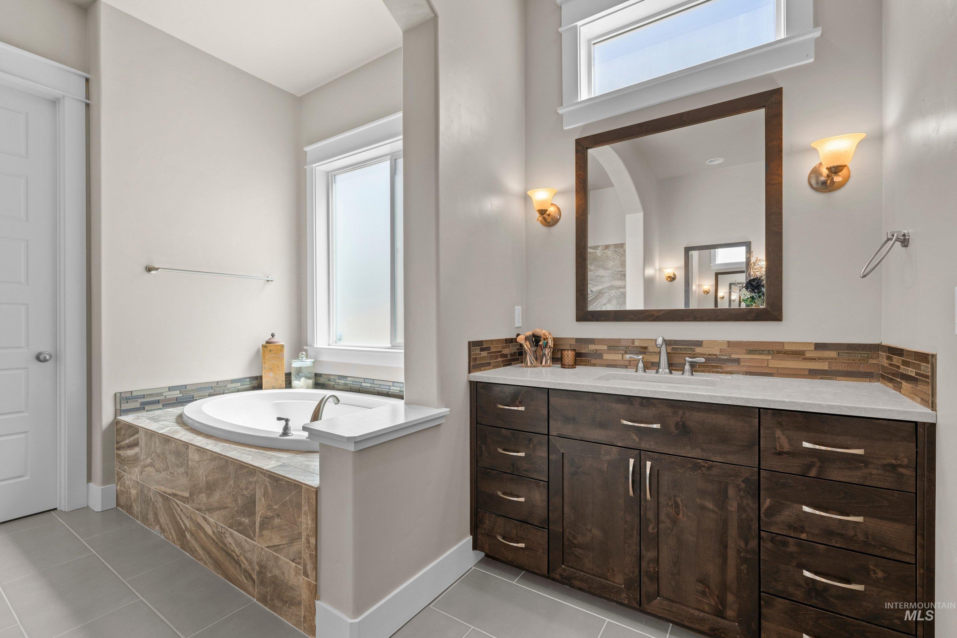 Bathroom featuring a built-in soaking tub with tile surround, a spacious vanity with a light-colored countertop, and a wood-framed mirror with sconce lighting