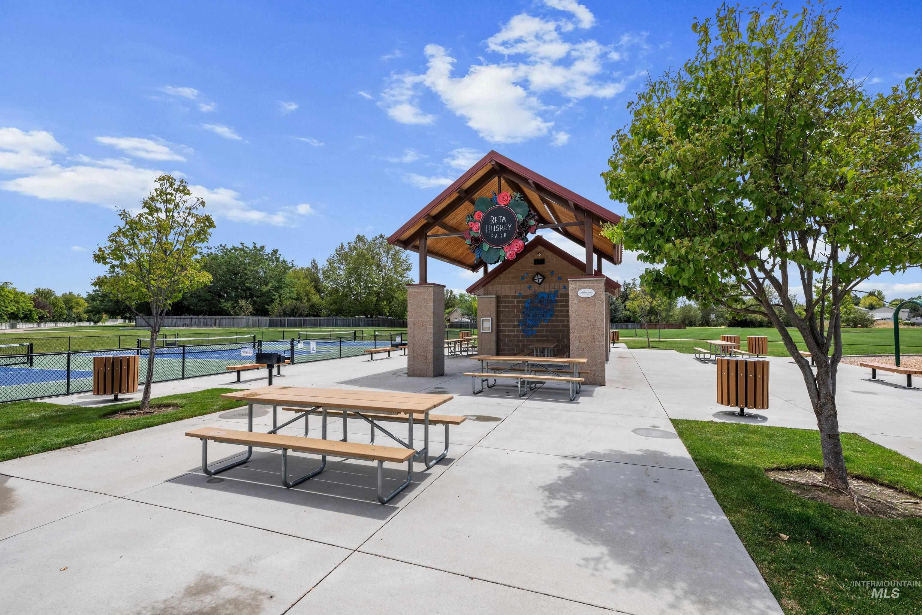 Park amenities featuring a covered picnic area with multiple picnic tables, a concrete pathway, a tennis court, and manicured green spaces