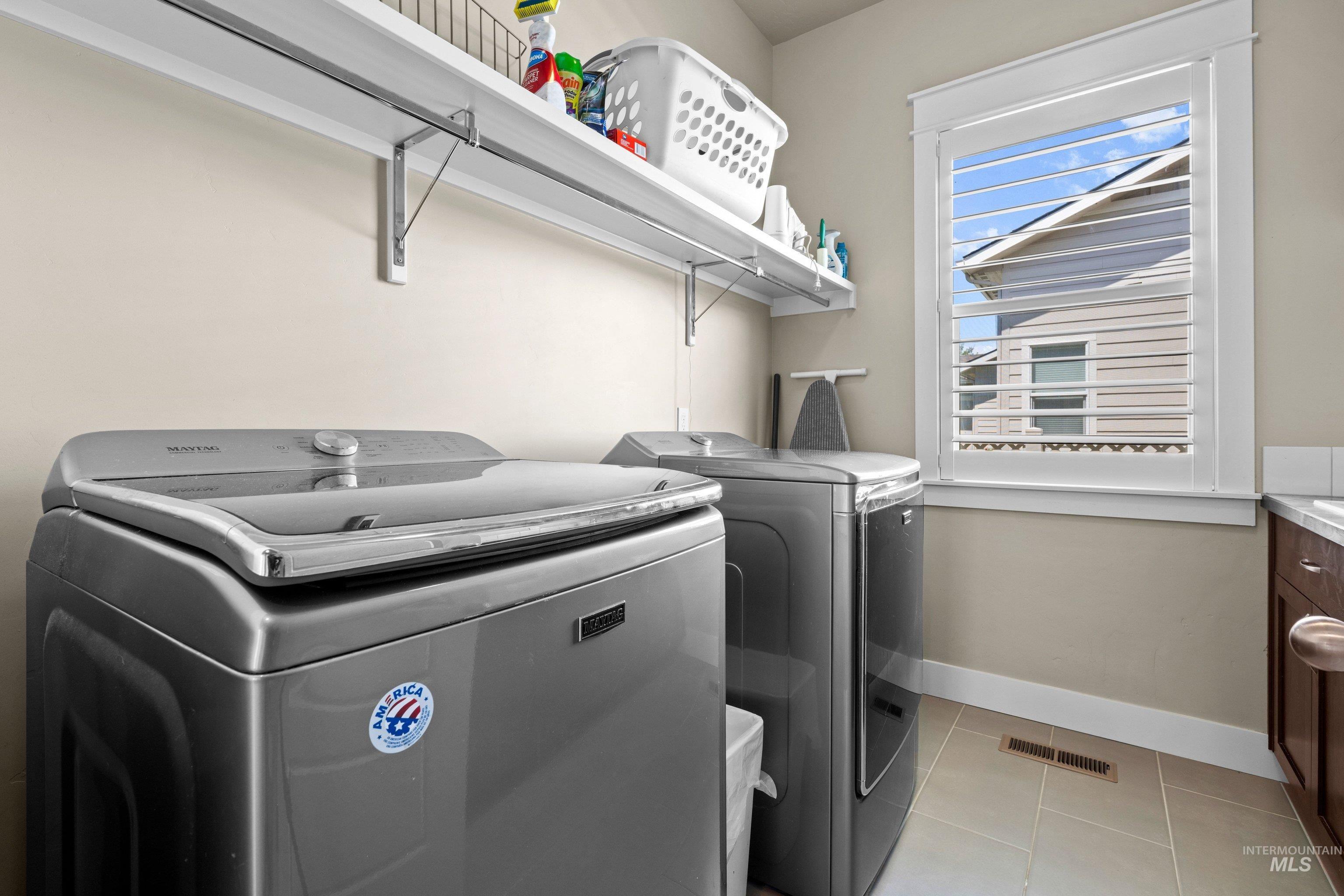 Dedicated laundry space featuring a window with white louvered shutters, overhead shelving, and tiled flooring