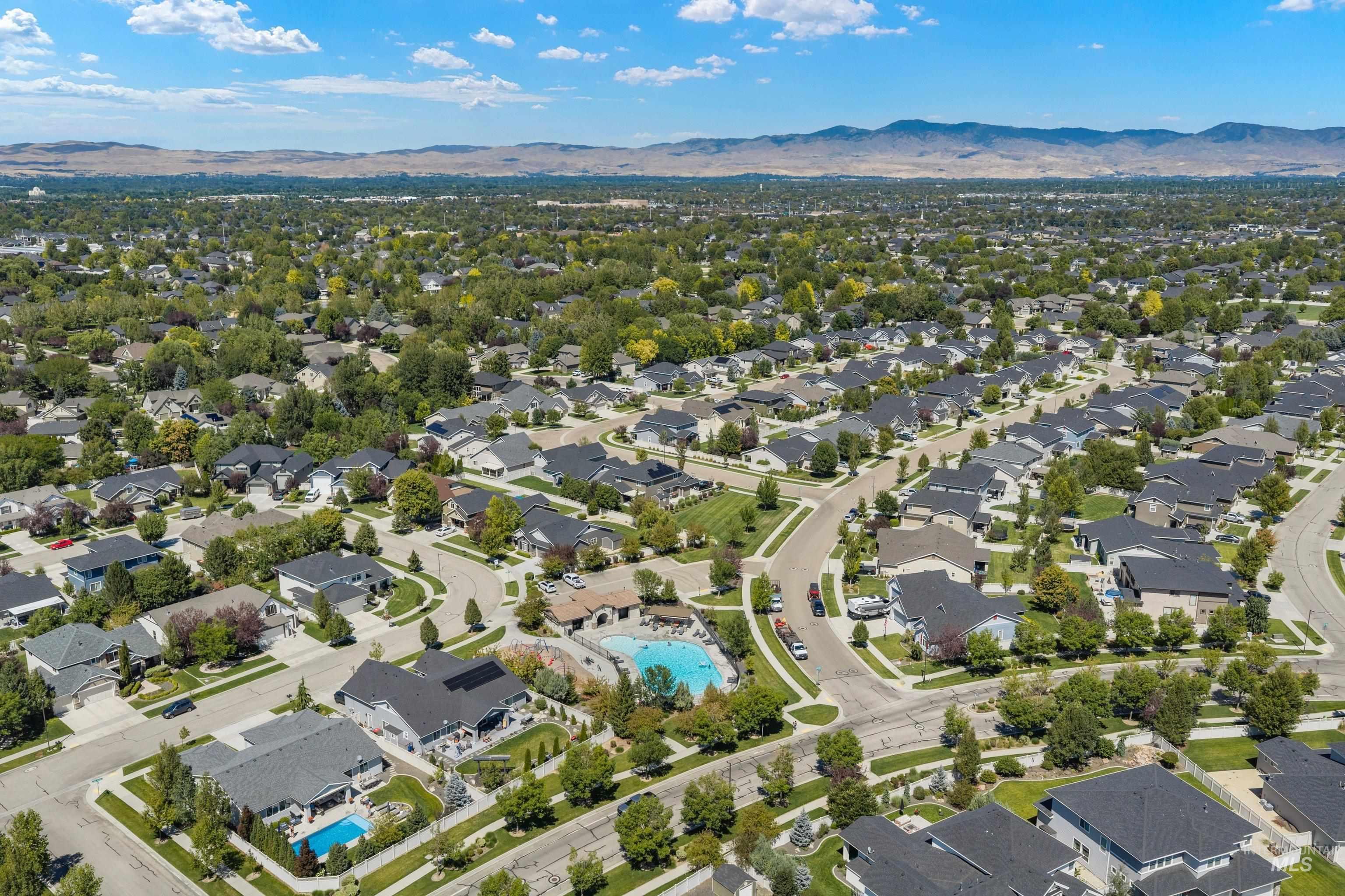 Aerial perspective of a residential community featuring numerous single-family homes with dark roofs, mature trees, winding streets, and a community pool with surrounding patio space