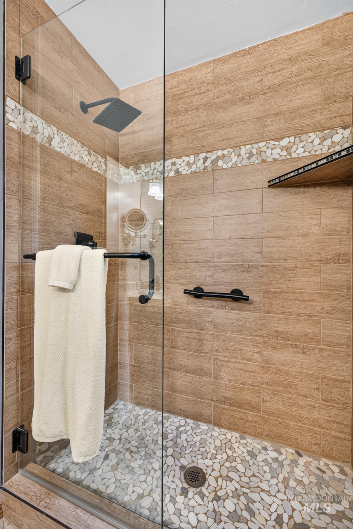 Walk-in shower featuring glass enclosure, wood-finish wall tiling with a mosaic pebble accent band, a pebble floor, matte black fixtures, and built-in shelving