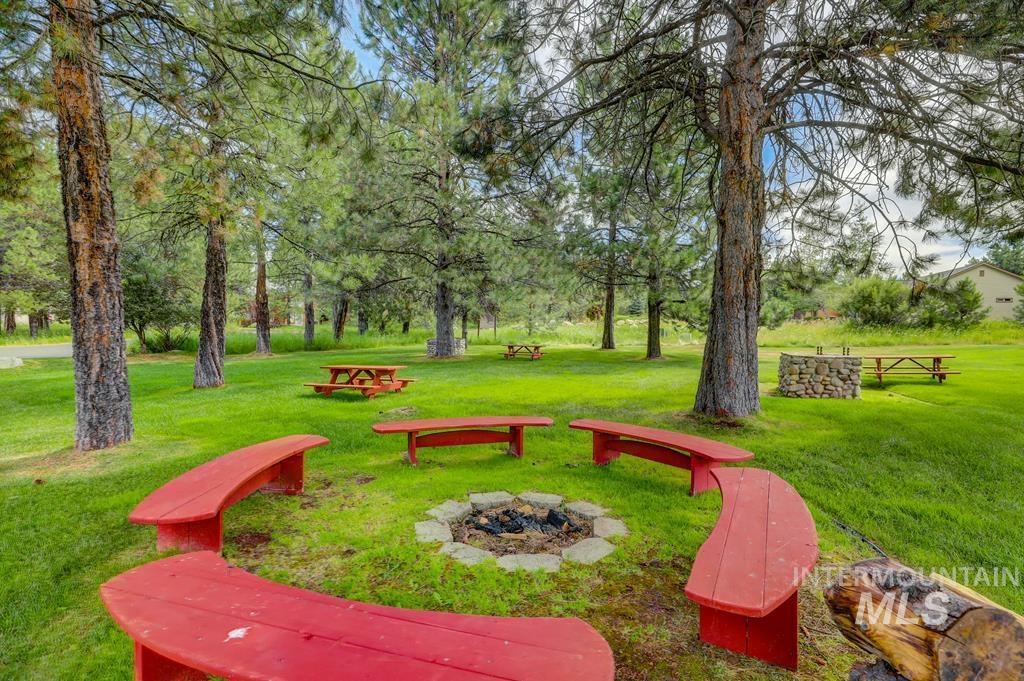 Expansive green space featuring a central fire pit surrounded by curved red benches, mature pine trees, and additional picnic tables