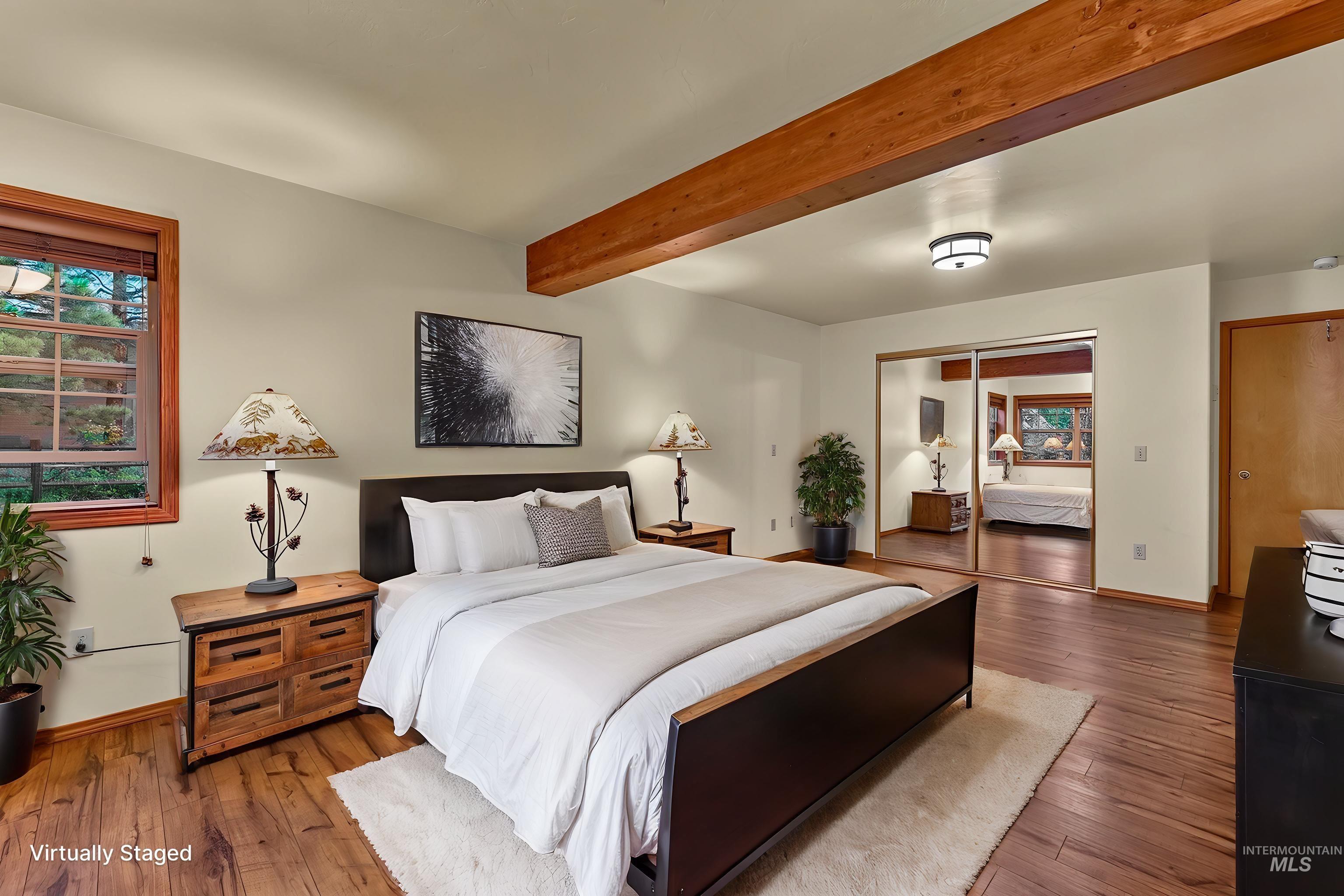 Spacious bedroom featuring wood-finish flooring, wood ceiling beams, a large window with wood trim, and mirrored closet doors