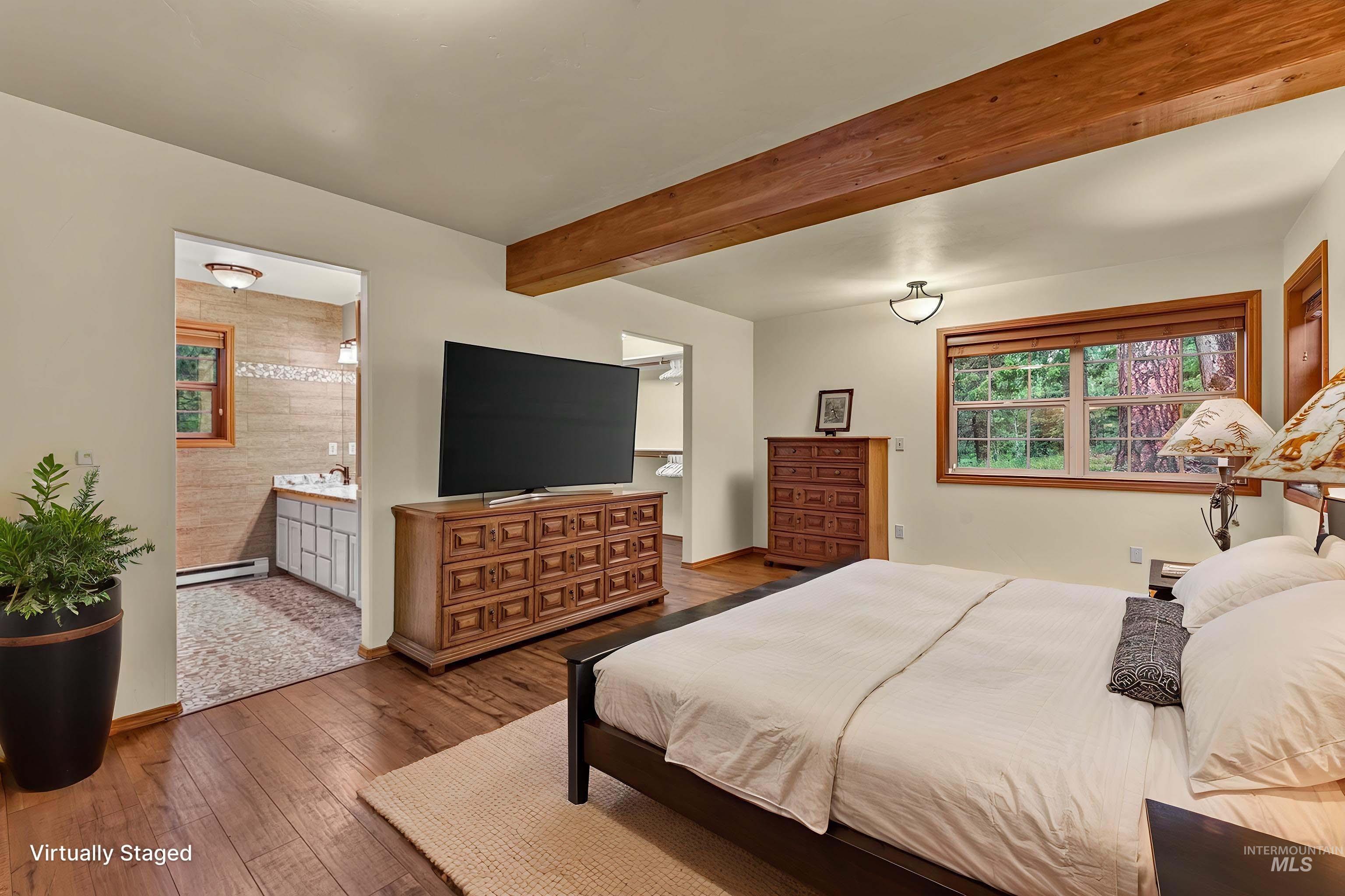 Spacious bedroom featuring wood-finish flooring, exposed wood beam, large window with natural wood trim, and a private en-suite bathroom with a vanity and tiled walls