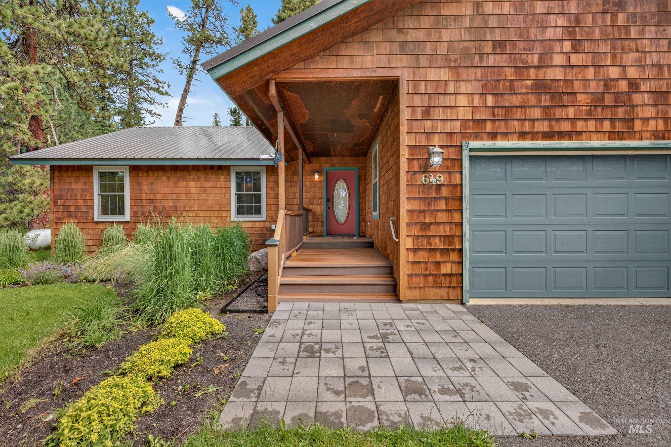 Cedar shake siding exterior with a red entry door, a gray paneled garage door, a covered porch, and a gray paver walkway