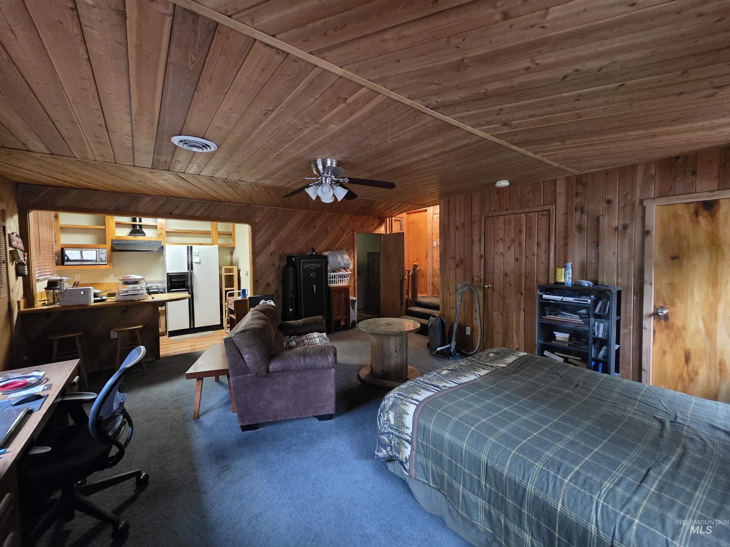 Bedroom with an office area, white refrigerator with ice dispenser, wood ceiling, dark colored carpet, and wood walls