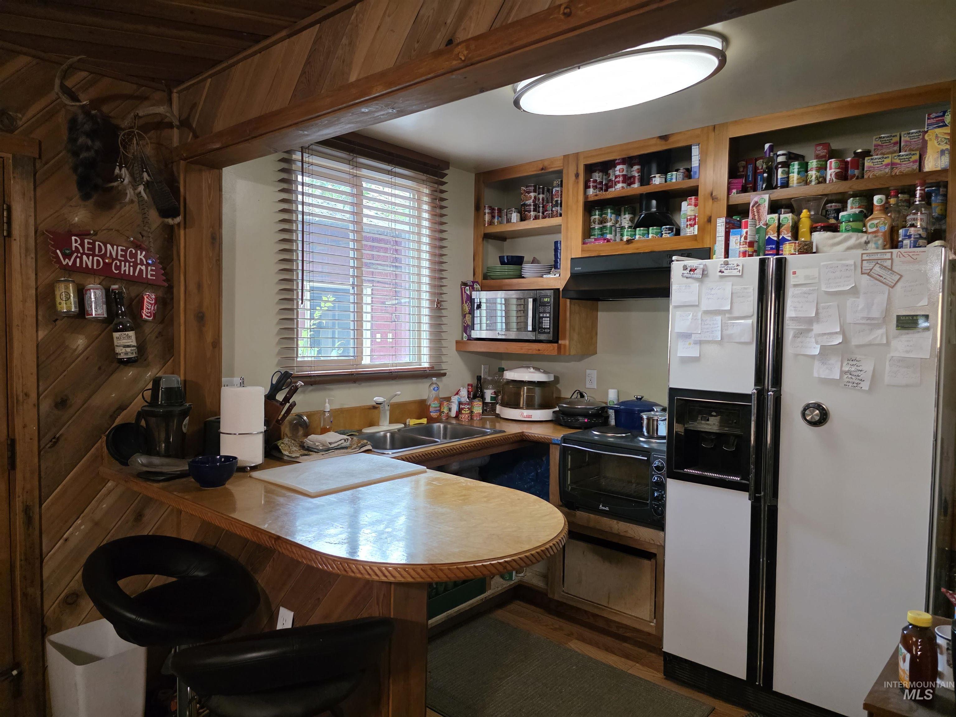 Kitchen with white refrigerator with ice dispenser, stainless steel microwave, a kitchen breakfast bar, light countertops, and under cabinet range hood