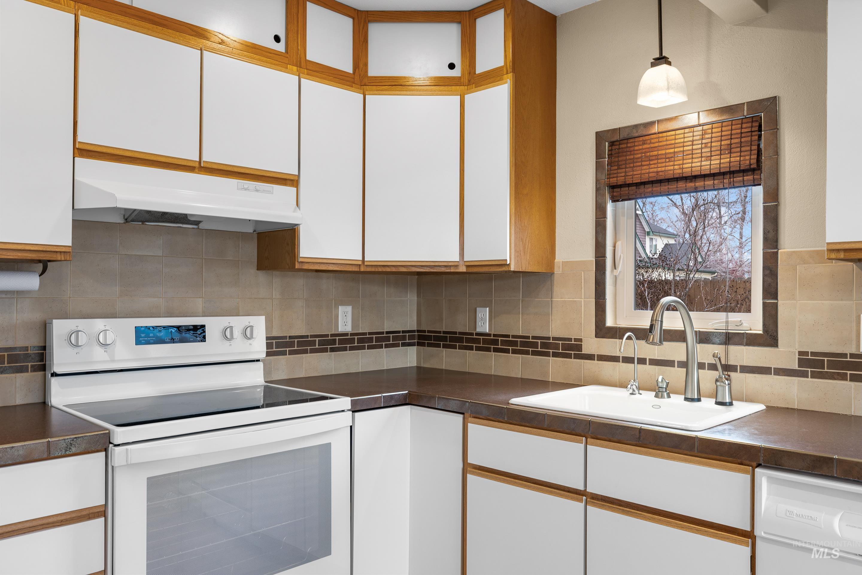 Kitchen with white appliances, two tone cabinets, dark countertops, hanging light fixtures, and decorative backsplash
