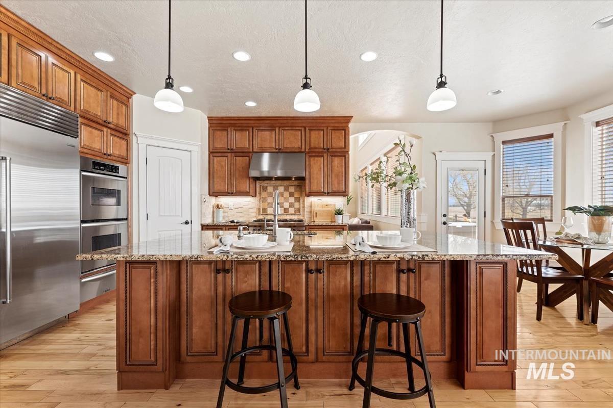 Kitchen with brown cabinetry, light stone counters, hanging light fixtures, tasteful backsplash, and appliances with stainless steel finishes