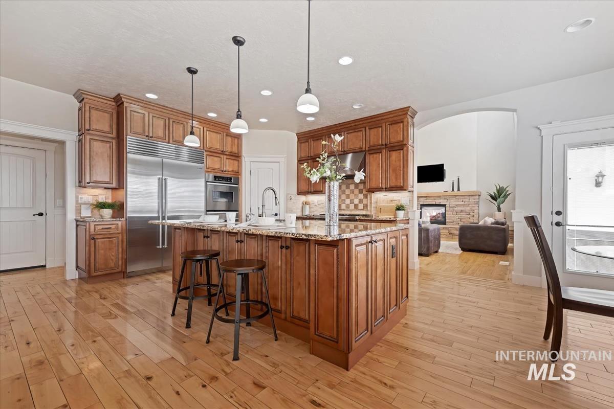 Kitchen with brown cabinetry, appliances with stainless steel finishes, a kitchen island with sink, light stone countertops, and recessed lighting