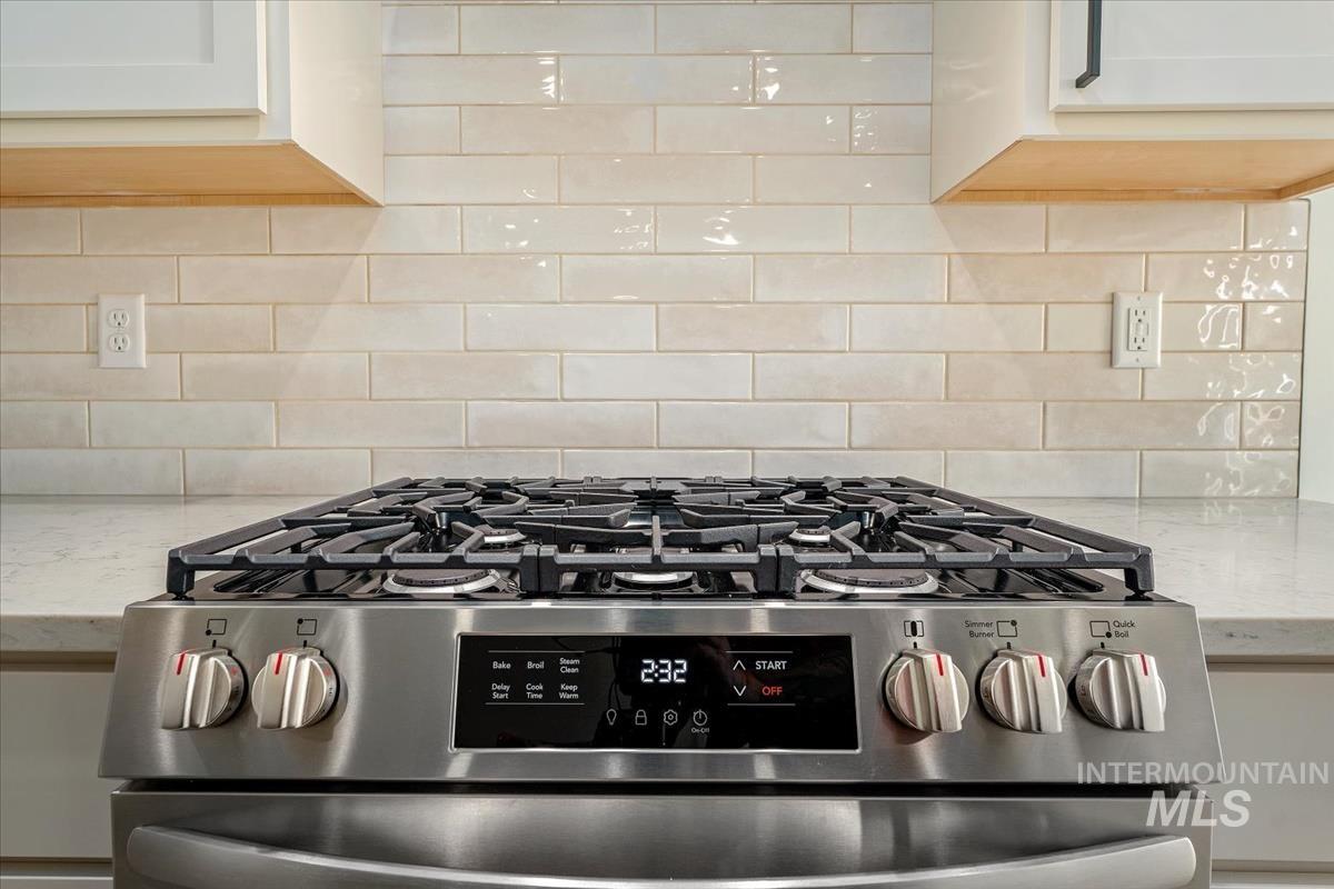 Kitchen view of stainless steel gas stove, backsplash, light stone counters, and white cabinets