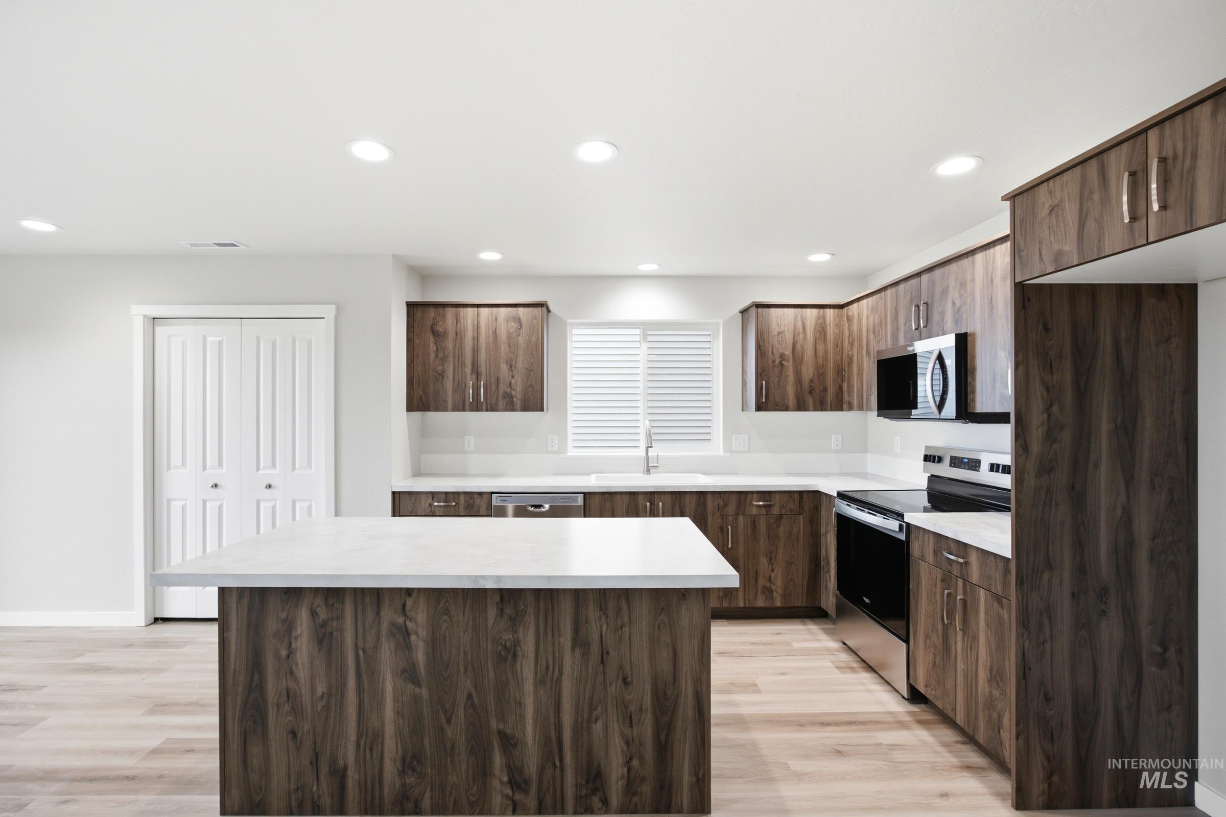 Contemporary kitchen featuring an expansive center island with white countertop, wood-finish cabinetry, stainless steel appliances, recessed lighting, and light wood-finish flooring