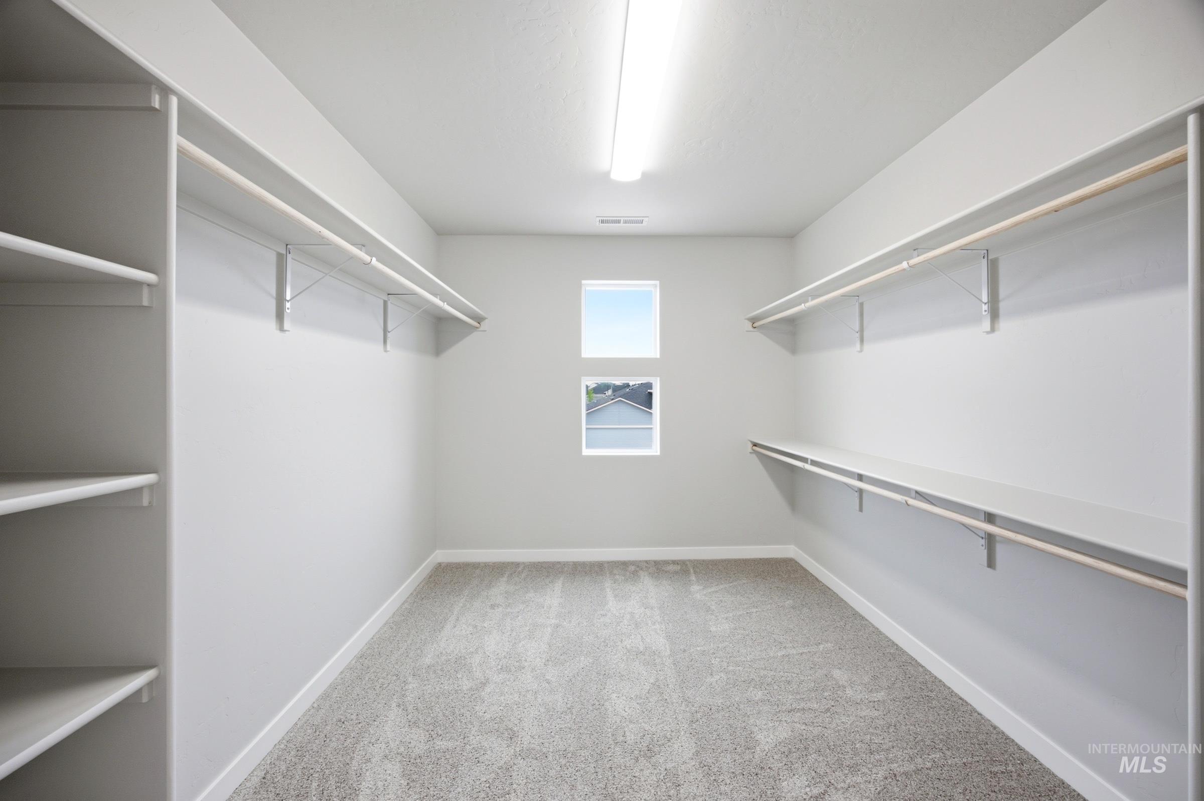Spacious walk-in closet featuring built-in shelving, dual hanging rods, light gray wall paint, white trim, and two square windows