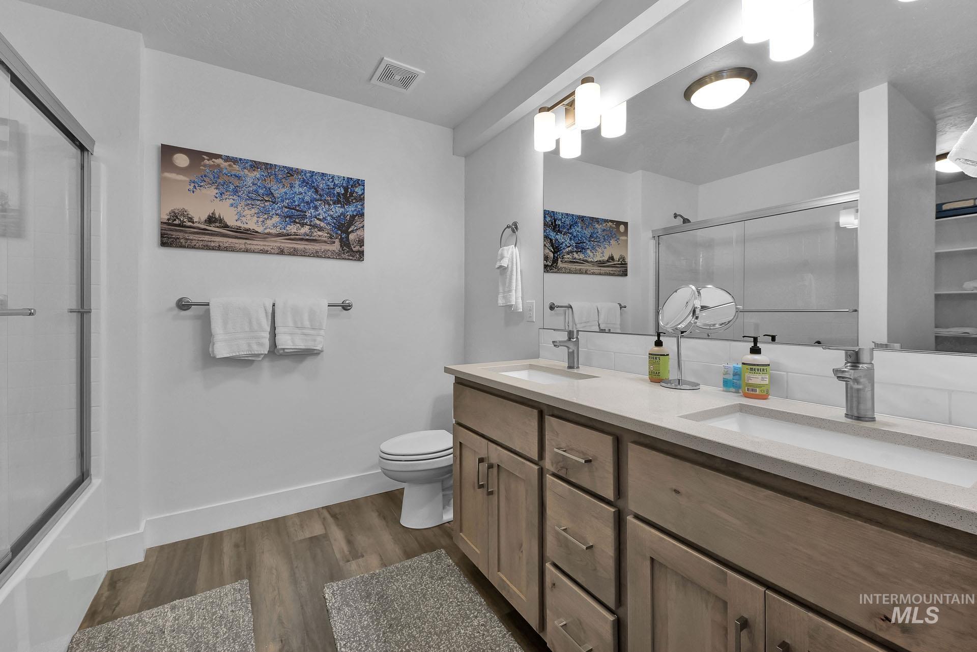Full bath featuring double vanity, a shower with shower door, and dark wood-style flooring