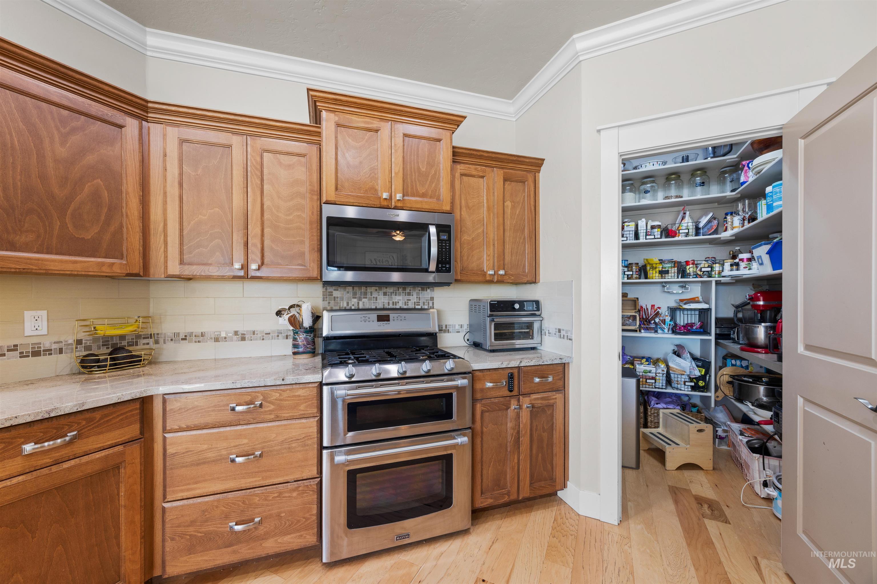 Kitchen with stainless steel appliances, ornamental molding, brown cabinets, light wood-style floors, and decorative backsplash