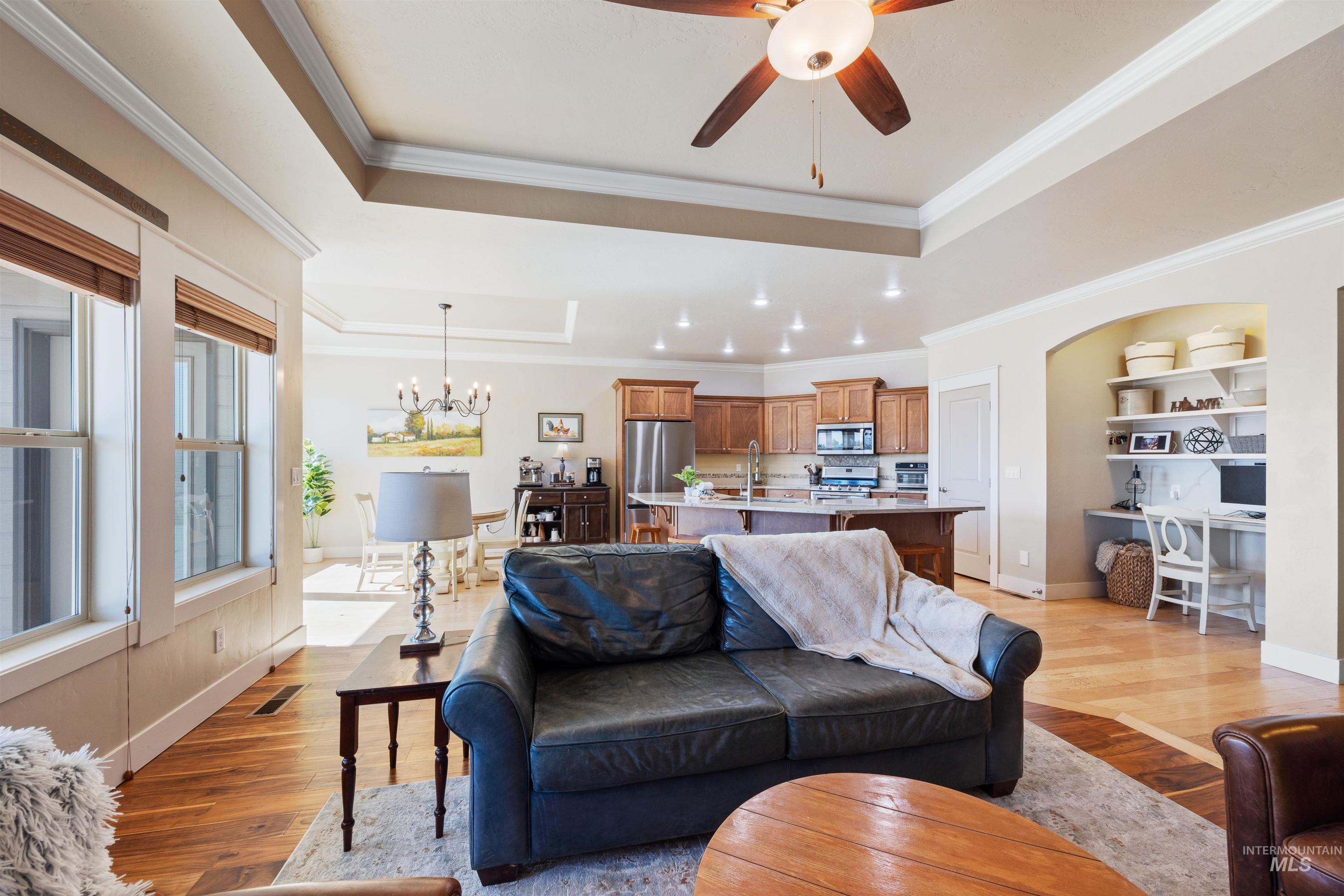 Living room featuring a raised ceiling, light wood-type flooring, ornamental molding, a chandelier, and a ceiling fan