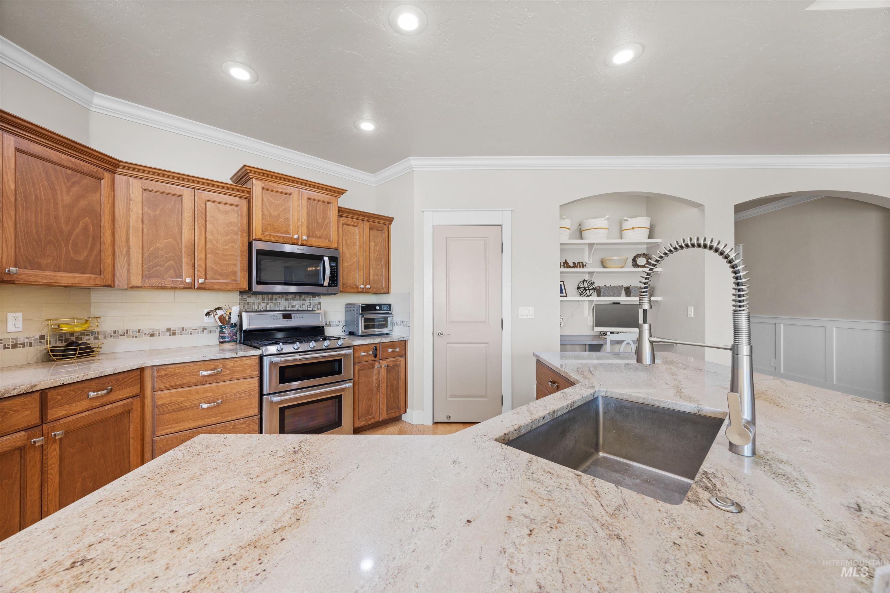 Kitchen with crown molding, brown cabinetry, appliances with stainless steel finishes, light stone counters, and built in features