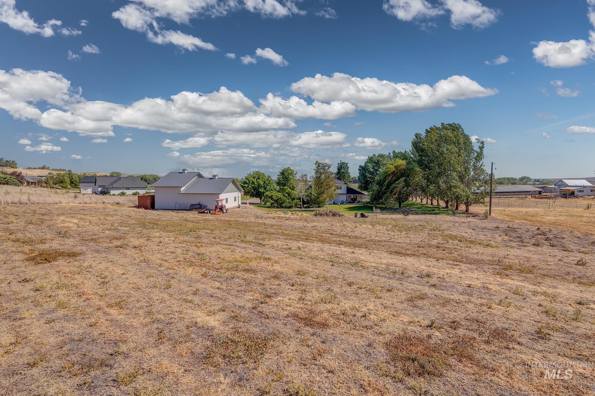 Expansive dryland acreage featuring scattered mature trees and distant residential structures under a bright blue sky with white clouds