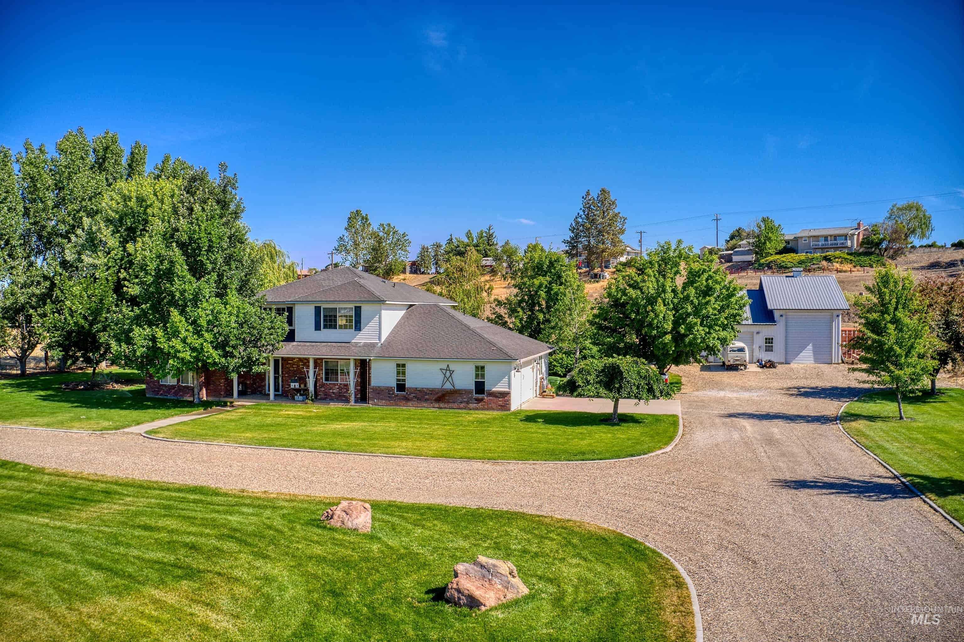 Spacious residential property featuring a gravel circular driveway, expansive green lawns, mature trees, a covered front porch with columns, and a detached auxiliary building with a metal roof