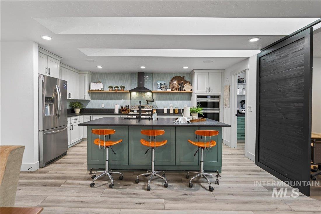Kitchen with open shelves, stainless steel appliances, wall chimney range hood, a kitchen bar, and a center island with sink