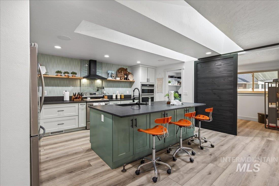 Kitchen featuring green cabinetry, open shelves, a kitchen bar, wall chimney exhaust hood, and decorative backsplash