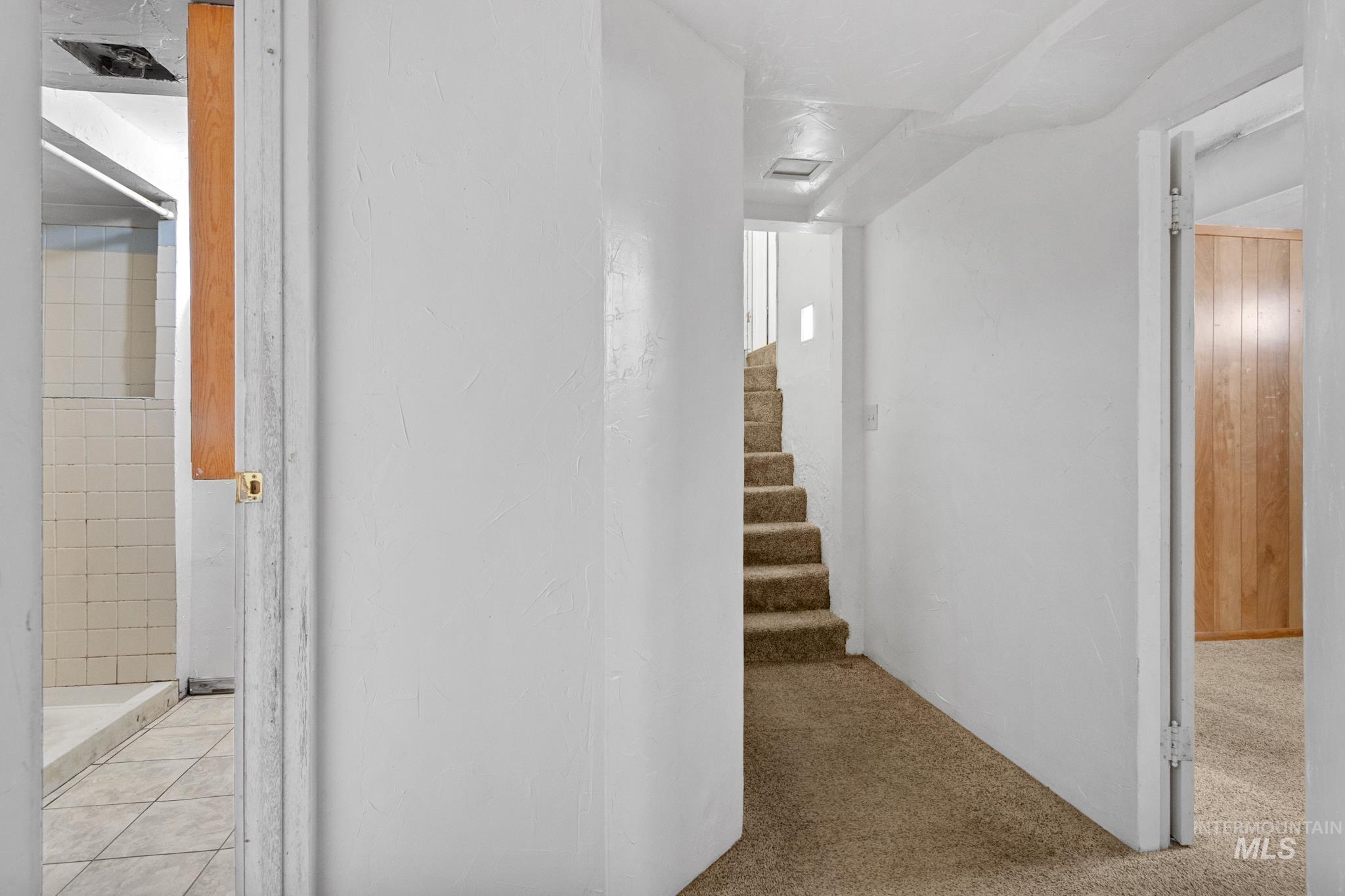 Carpeted hallway featuring a staircase, white painted walls, and two doorways revealing additional interior spaces