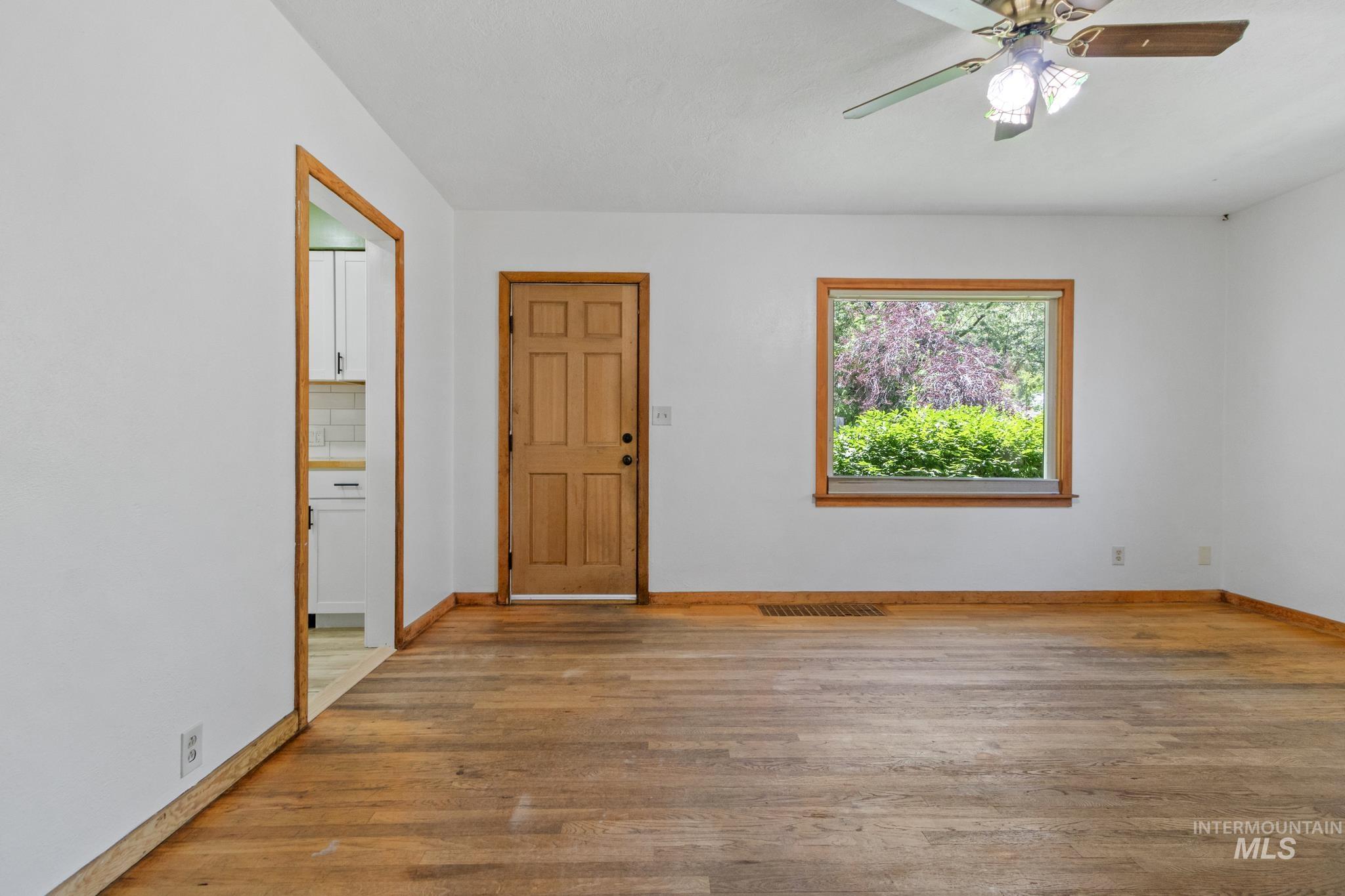 Living space featuring wood-finish flooring, a wood-framed window with exterior foliage, a wood paneled door, and a ceiling fan with integrated lighting