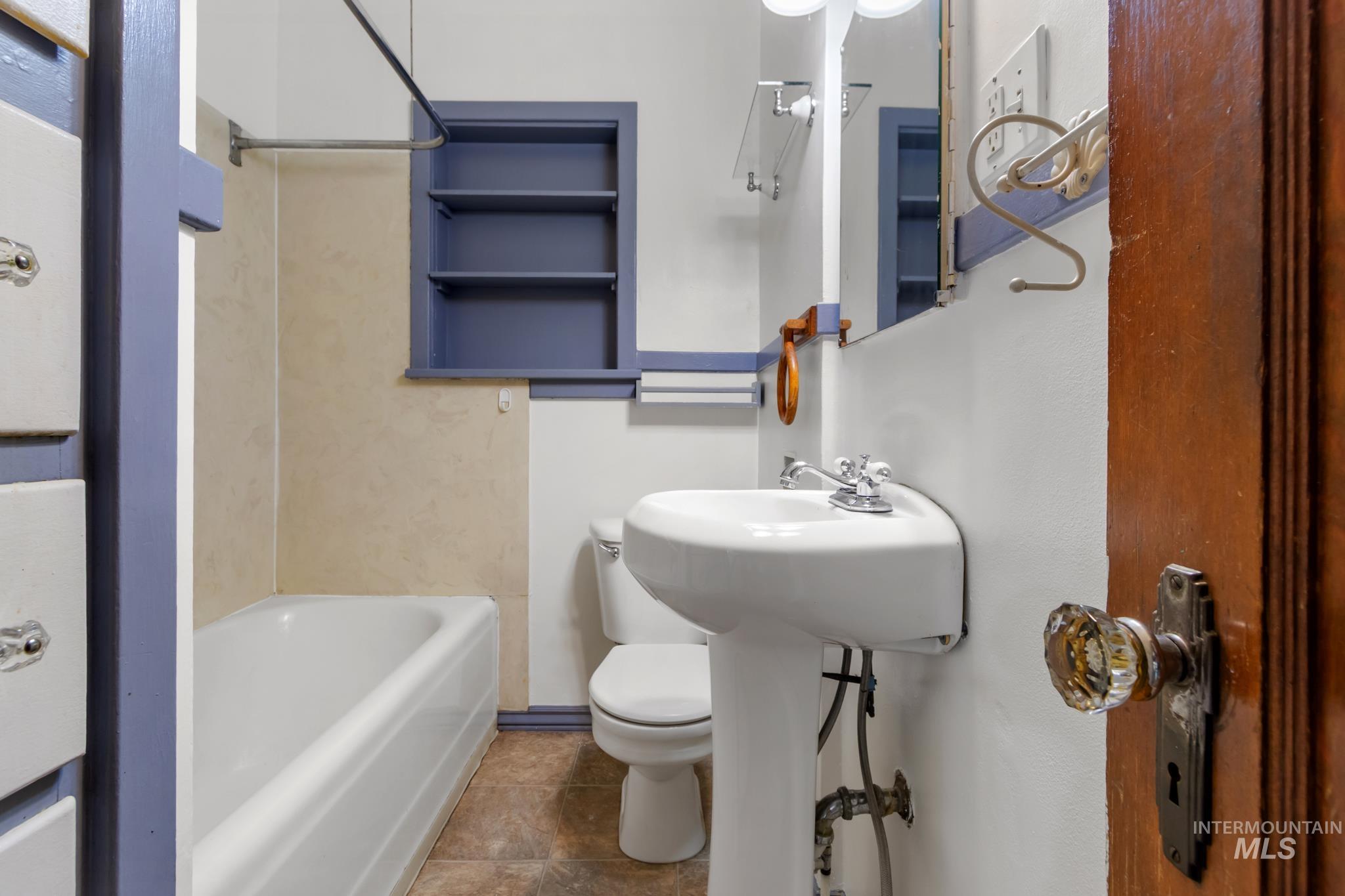 Pedestal sink with chrome fixtures, white toilet, built-in tub with tile surround, built-in shelving, and wood-finish door with crystal knob