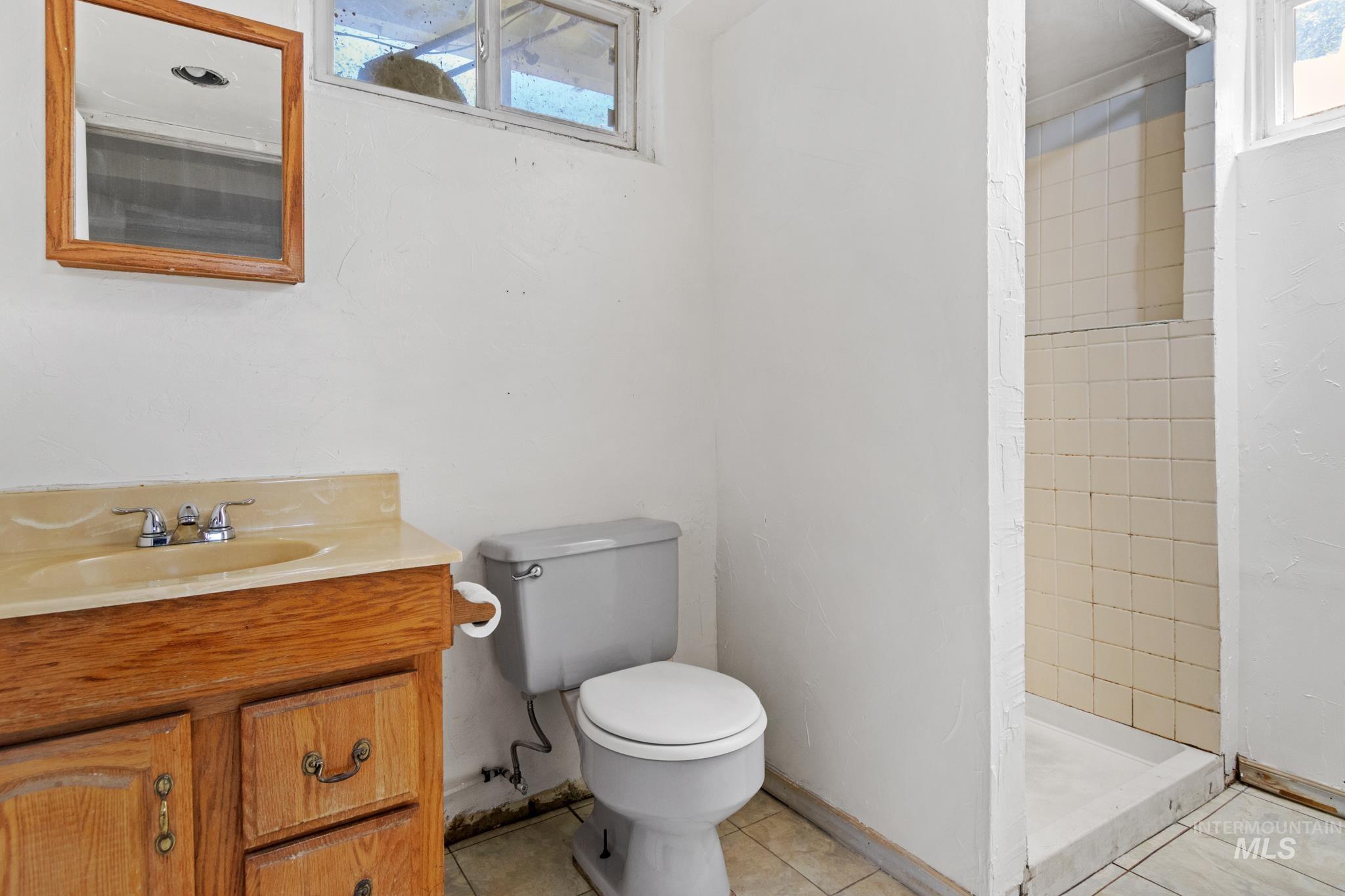 Bathroom featuring a wood vanity with integrated sink, a commode, a tiled shower stall, and a window