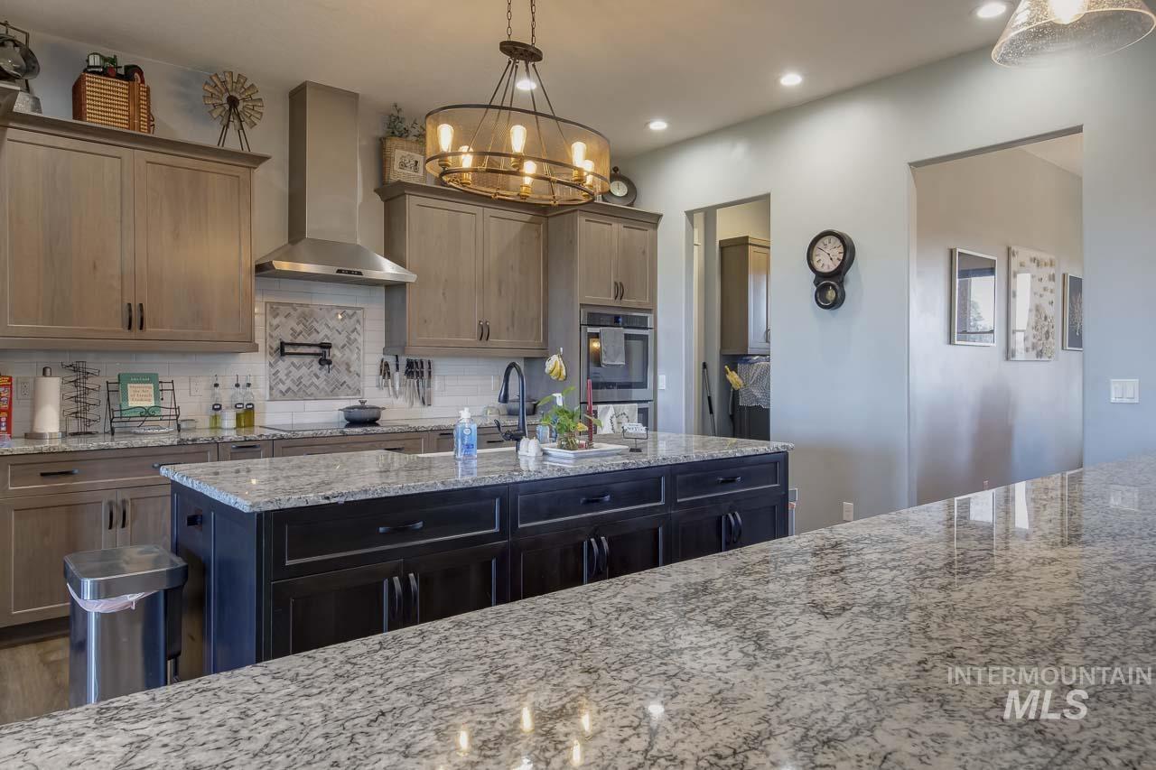 Kitchen with wall chimney range hood, light stone counters, backsplash, a chandelier, and decorative light fixtures
