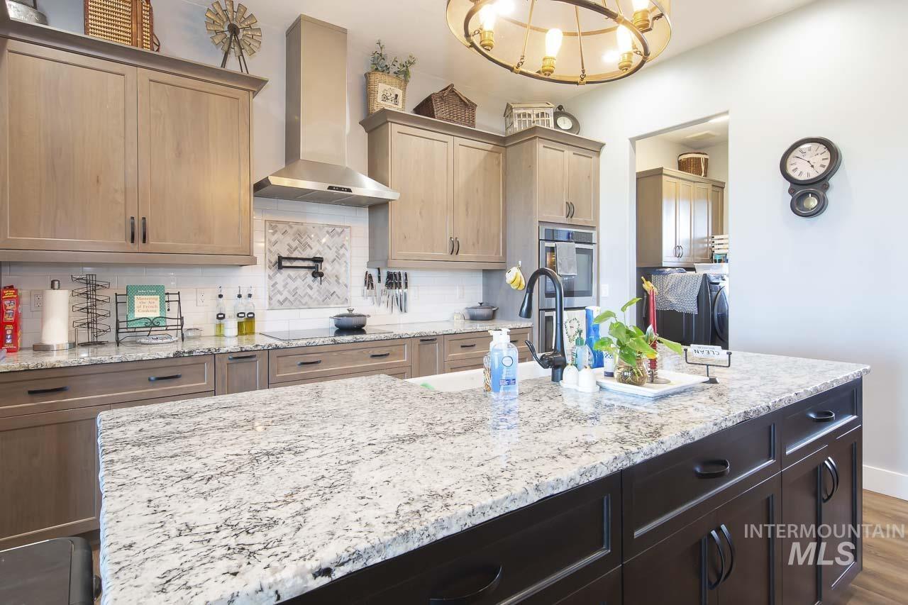 Kitchen featuring wall chimney exhaust hood, tasteful backsplash, light stone counters, dark wood finished floors, and a chandelier
