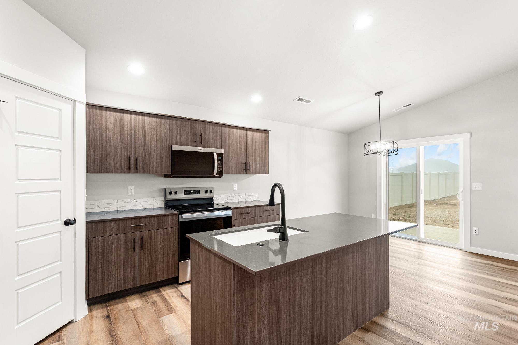 Kitchen featuring appliances with stainless steel finishes, light wood-style floors, pendant lighting, a center island with sink, and dark stone countertops