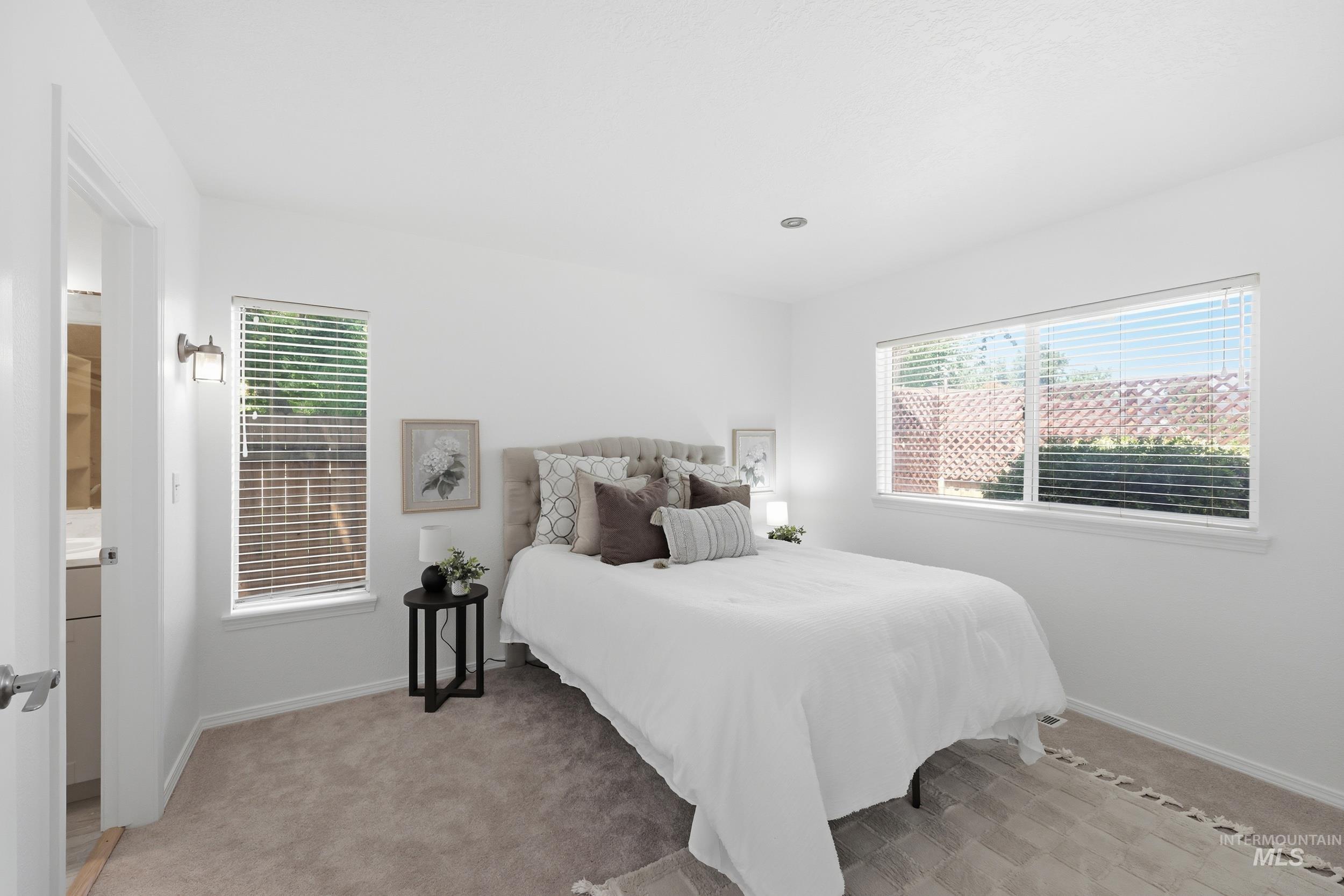 Bright bedroom featuring two windows with blinds, a recessed ceiling light, wall-mounted sconce, and neutral carpeting