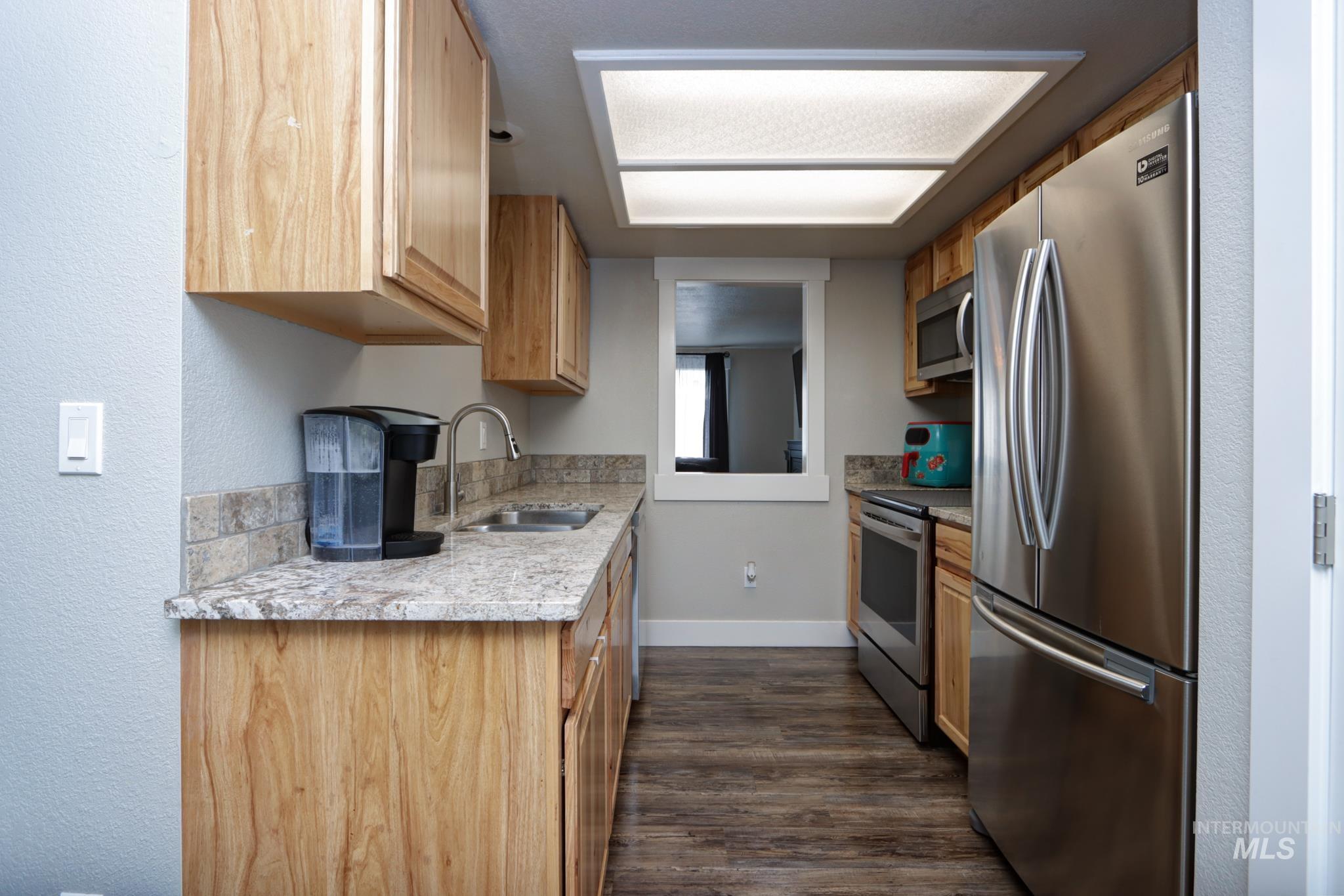 Kitchen featuring stainless steel appliances, dark wood-type flooring, a textured wall, light wood finish cabinetry, and light stone countertops