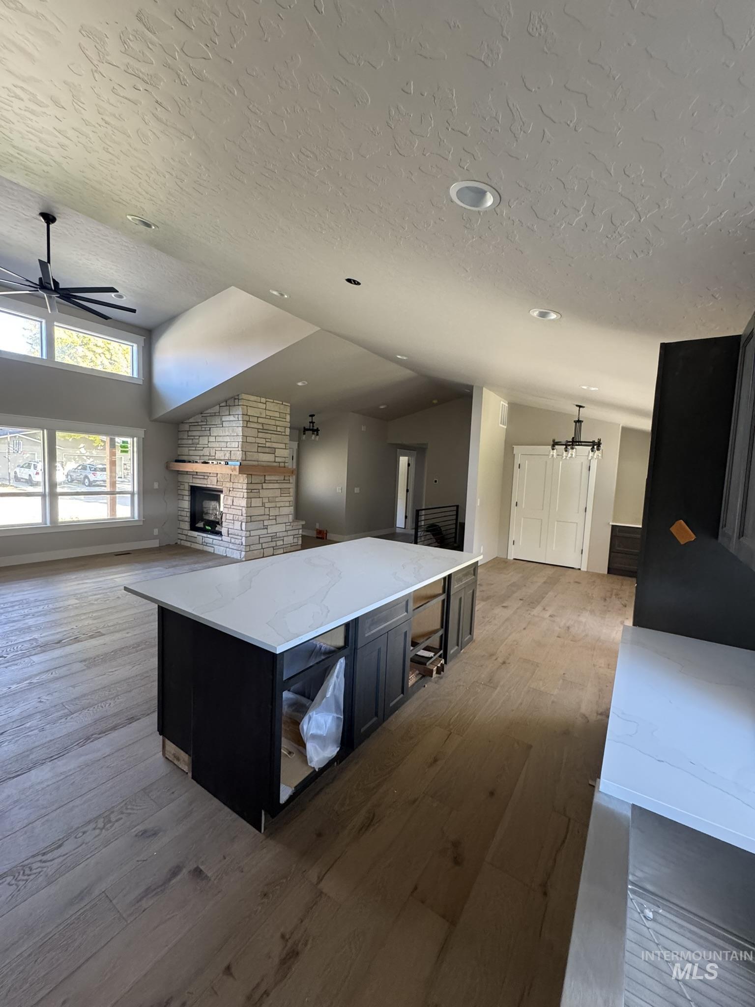 Kitchen featuring lofted ceiling, dark cabinetry, light stone countertops, wood finished floors, and a stone fireplace
