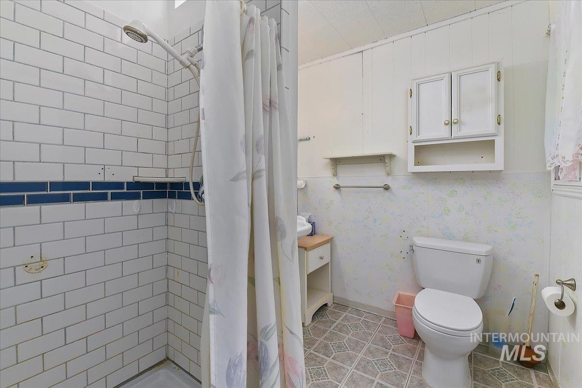 Shower with white subway tile and blue accent band, white wood paneling, wall-mounted medicine cabinet, white toilet, and patterned floor tile