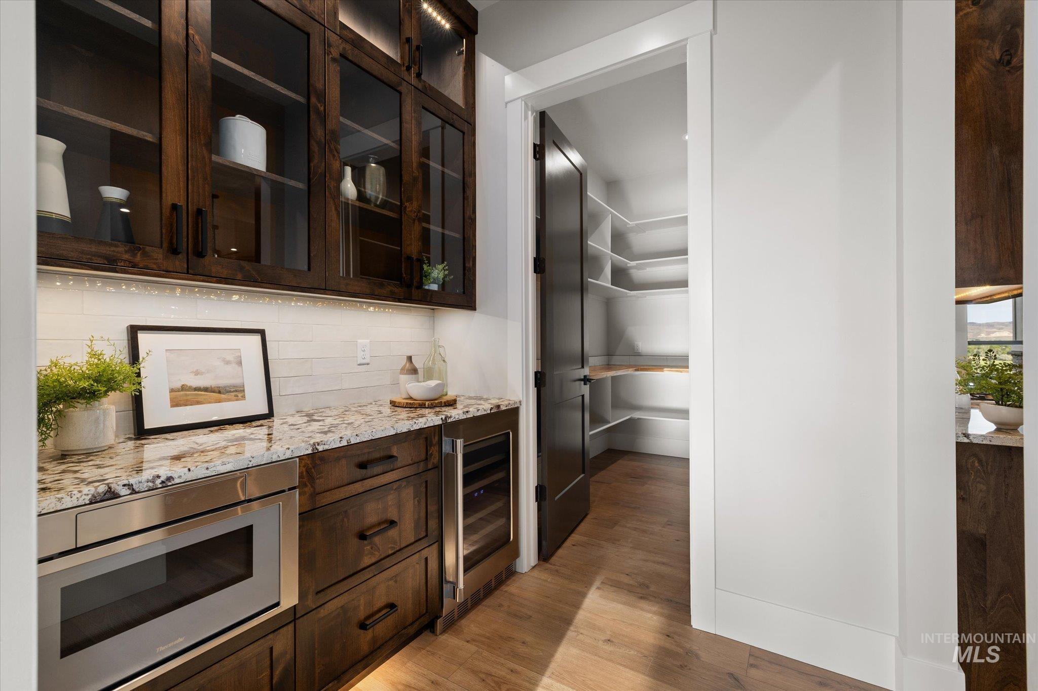 Bar with dark brown cabinetry, decorative backsplash, light wood-style flooring, light stone counters, and stainless steel microwave
