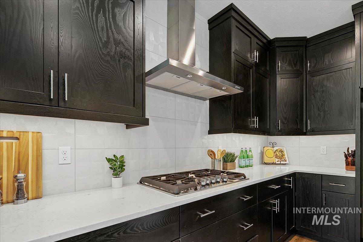 Kitchen with decorative backsplash, wall chimney exhaust hood, dark cabinetry, and light stone countertops