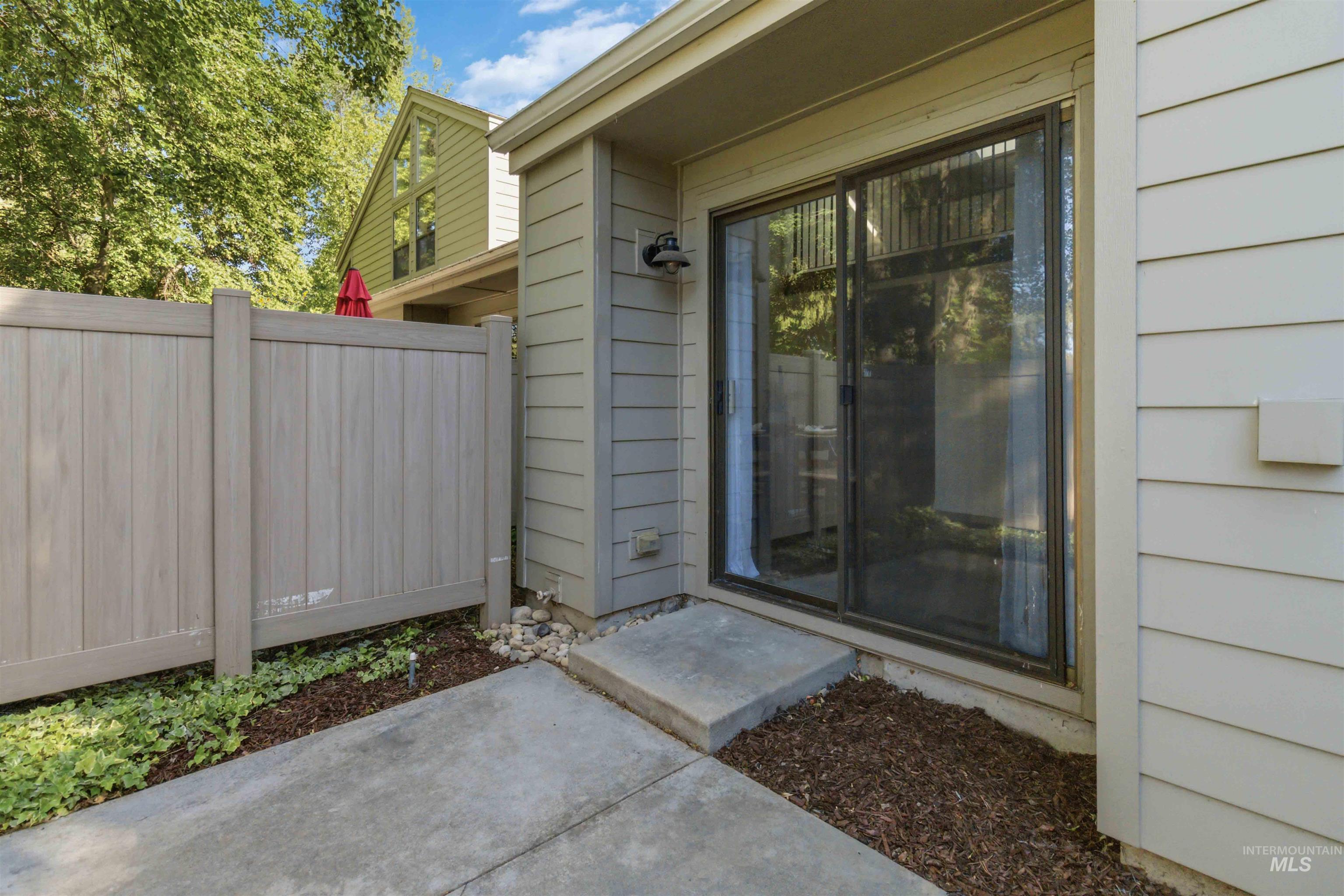 Private patio area featuring a sliding glass door entry, concrete pad, horizontal panel fencing, exterior wall lantern, and wood chip landscaping