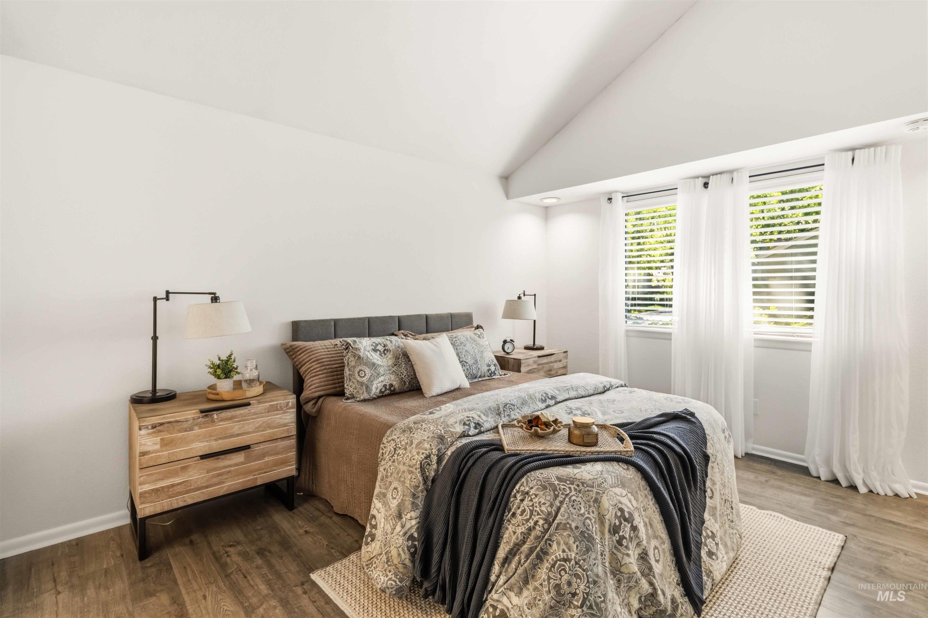 Vaulted ceiling bedroom featuring wood-finish flooring, two window openings with blinds, a gray upholstered headboard, and recessed lighting