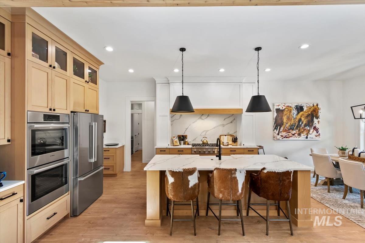 Kitchen featuring light wood-type flooring, decorative light fixtures, appliances with stainless steel finishes, a kitchen island with sink, and tasteful backsplash