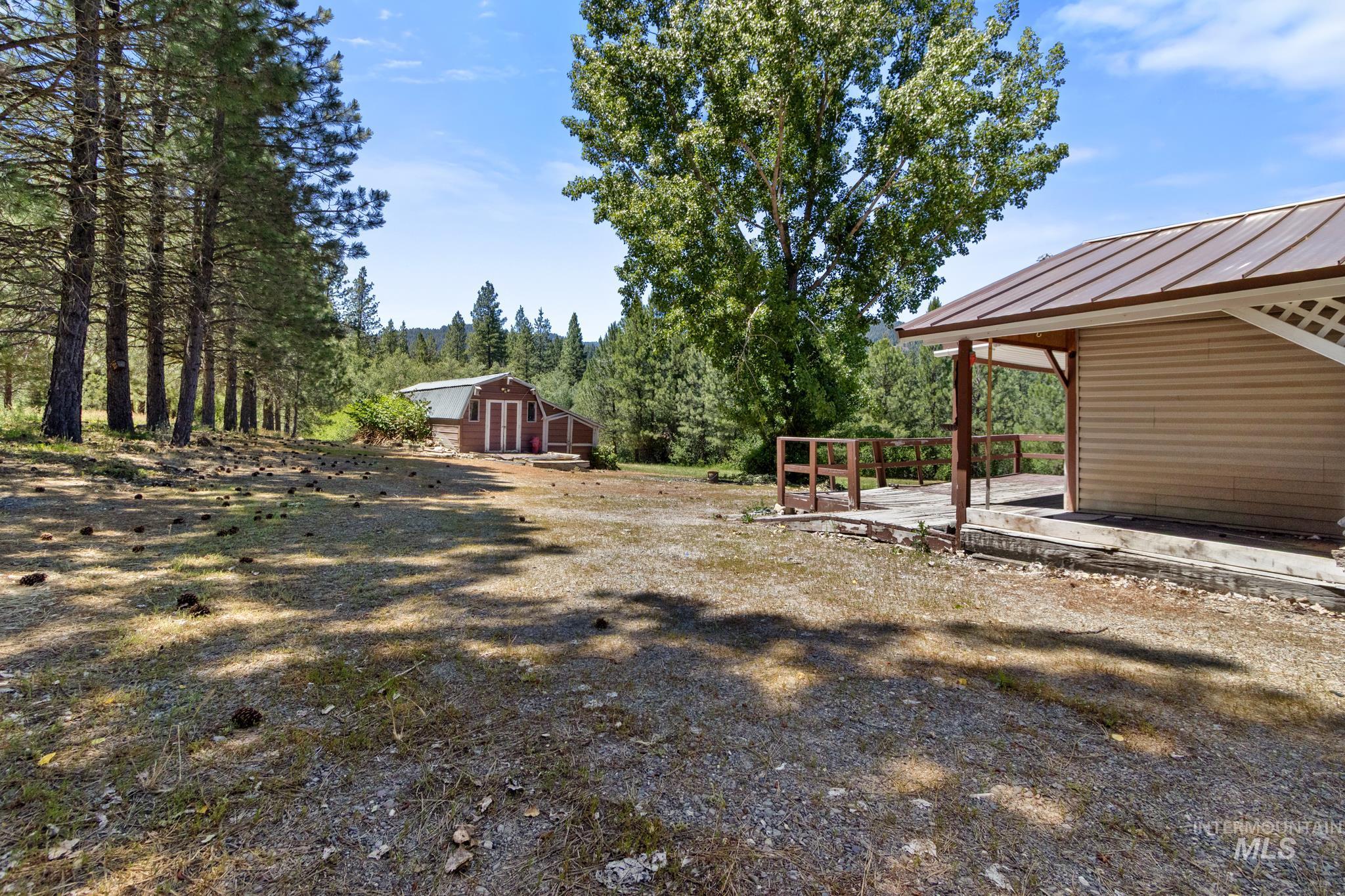 Expansive outdoor space featuring a covered porch with a metal roof and lattice detailing, a red barn structure, mature trees, and a gravel and grass ground cover