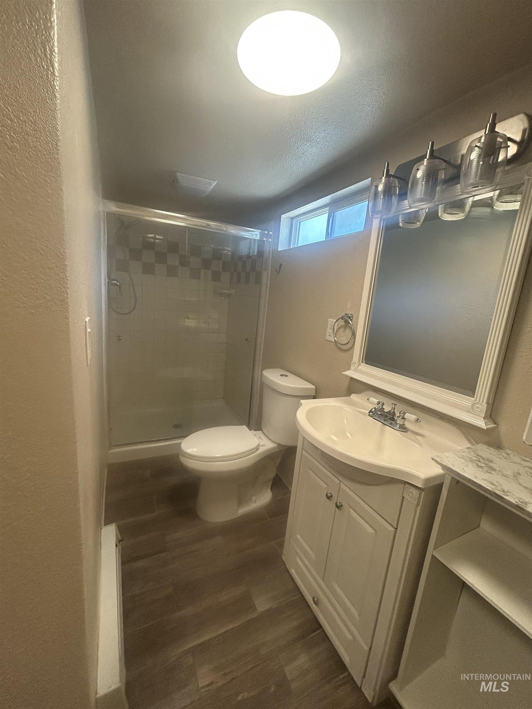Bathroom featuring a glass-enclosed shower, wood-finish flooring, a white vanity with integrated sink, a framed mirror, and an overhead light fixture
