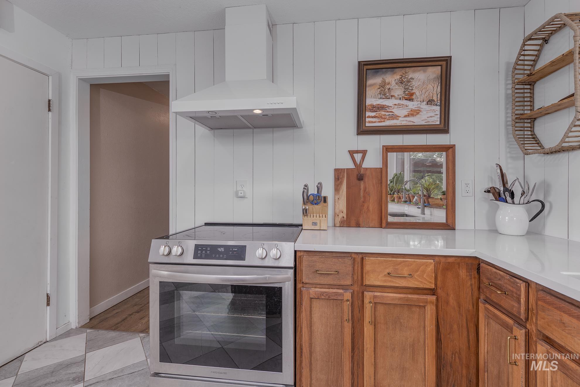 Kitchen featuring electric range, wall chimney exhaust hood, brown cabinets, and light stone counters