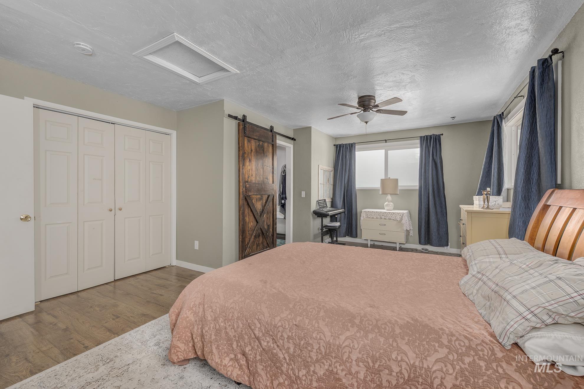 Bedroom featuring attic access, a textured ceiling, wood finished floors, a barn door, and ceiling fan