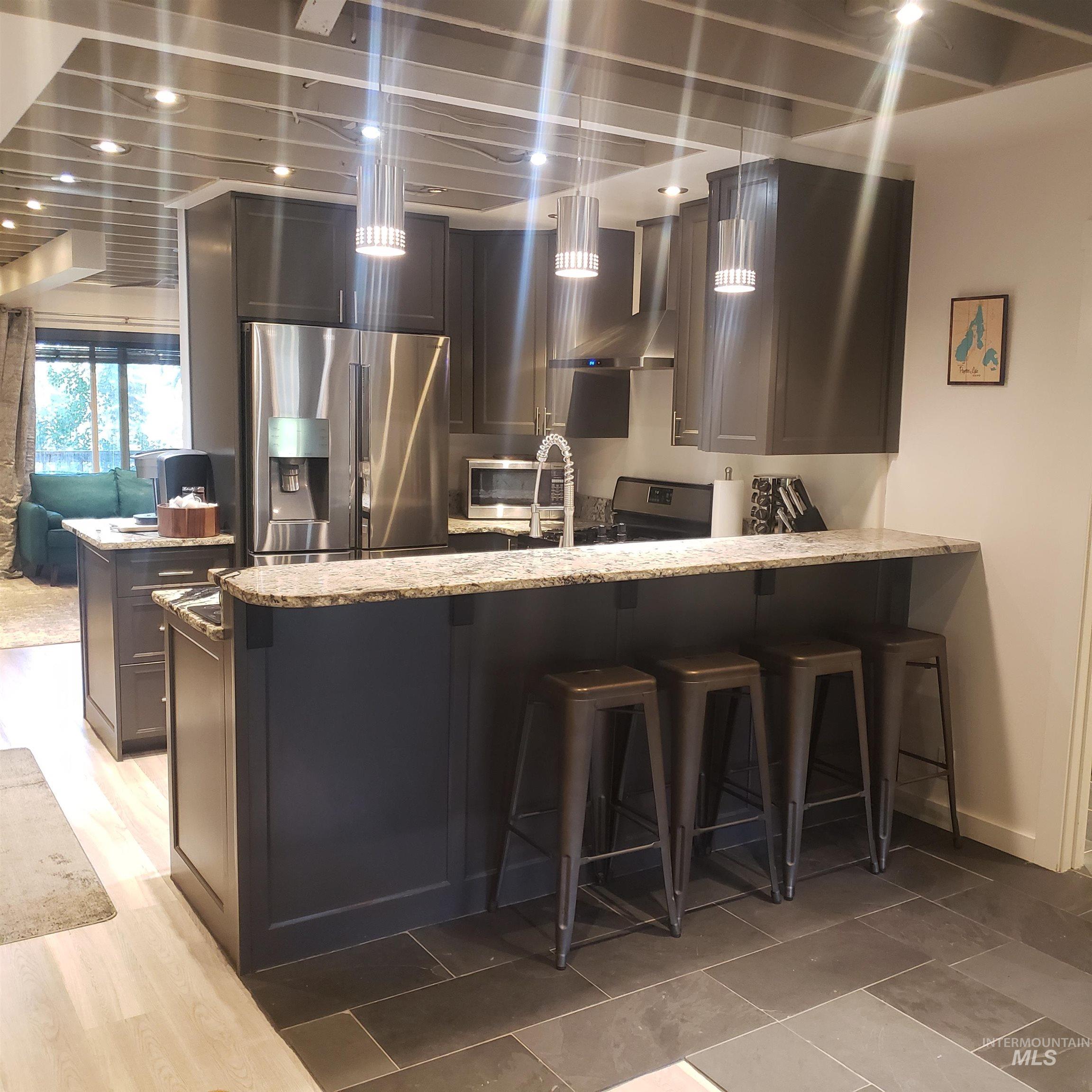 Kitchen featuring a breakfast bar, stainless steel appliances, light stone countertops, decorative light fixtures, and wall chimney range hood