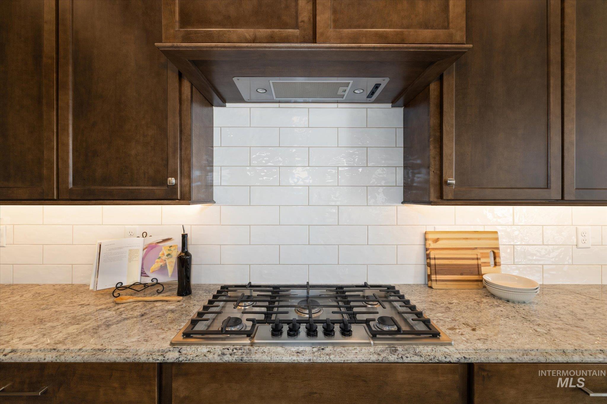 Kitchen with ventilation hood, light stone counters, stainless steel gas cooktop, and dark brown cabinets
